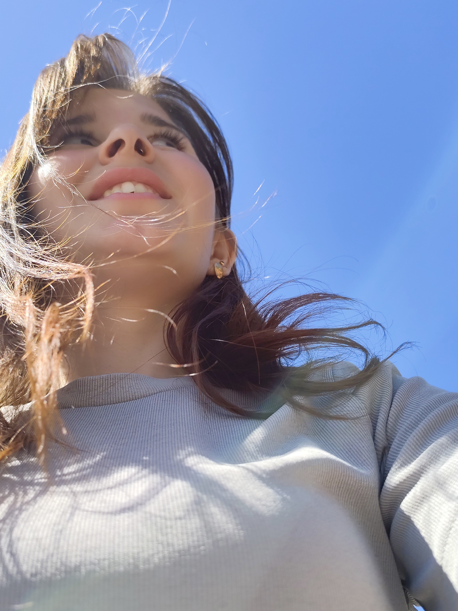 A young woman is looking up into a clear blue sky. Her brown hair is blowing in the wind, and she is wearing a grey sweater. Her location is unknown.