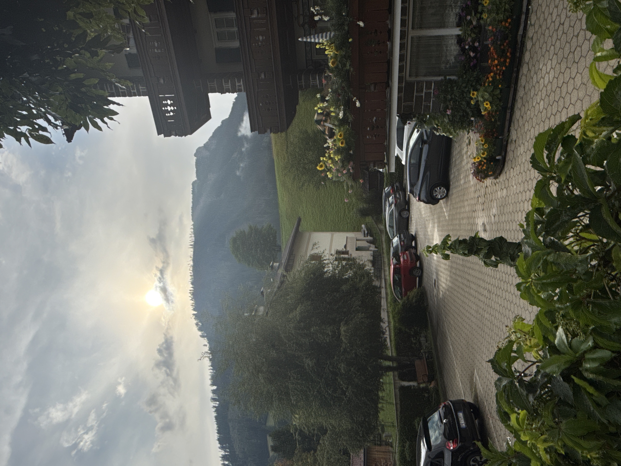 A view of a parking lot and a building in Moena, Italy. The sun is peeking through cloudy skies, casting a soft light on a tree-covered hillside in the background. In the foreground, a paved parking lot is visible, with several cars parked. A multi-story building with balconies and window shutters stands to the right of the parking lot. Flowers adorn a planter box on a balcony. The overall scene is tranquil and hints at a mountainous region.