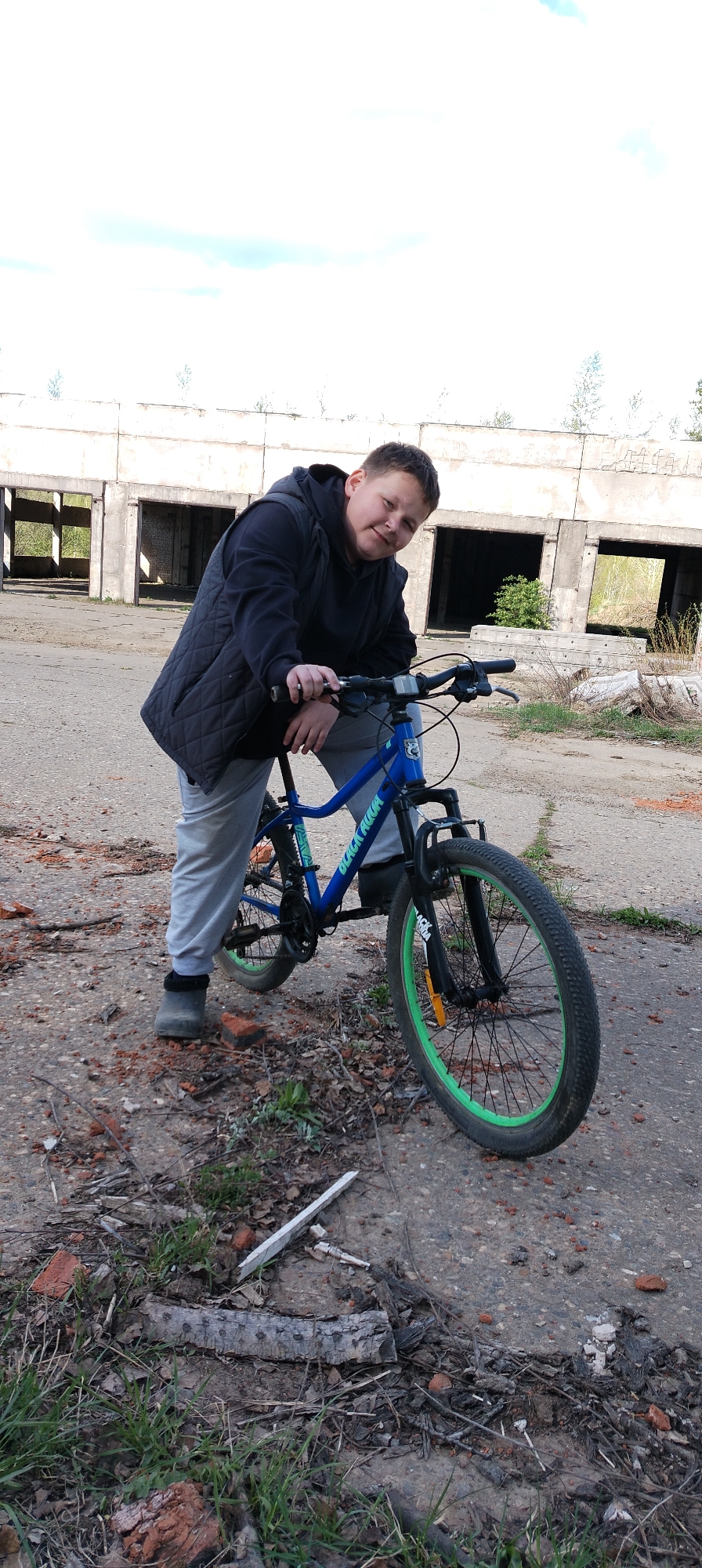 A young person is posing with a blue and black mountain bike with green rims in Зипуново, Russia. The bike is in the foreground. The person is leaning on the bike's handlebars. The background includes a dilapidated, concrete structure with open doorways. The ground is a mix of dirt, concrete, grass, and scattered debris including bricks and wood. The sky is overcast.