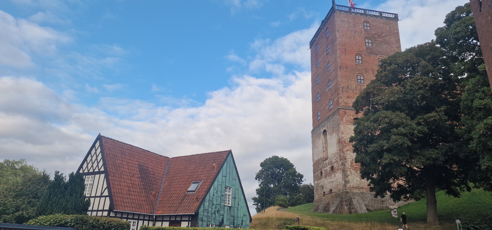 The image shows the Koldinghus castle in Kolding, Denmark. The castle is a tall, brick tower with a red and white flag at the top. To the left of the tower is a traditional Danish house with a red tiled roof and white half-timbered walls. The house is nestled among trees and greenery. A person can be seen walking on the grassy slope in front of the tower, while another person stands near the base of the tower. The sky is blue with scattered white clouds. The scene appears to be set on a sunny day.