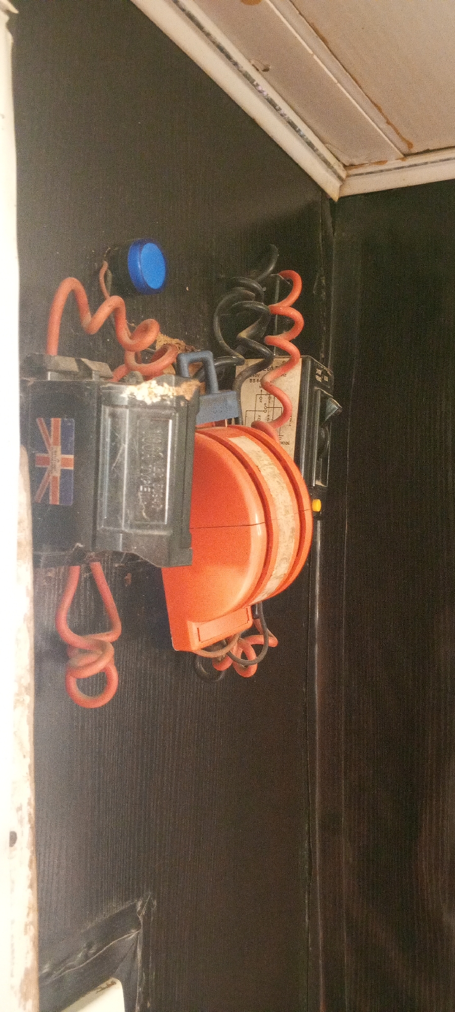 The image shows a close-up, side-view of electrical wiring and components mounted on a dark brown, wood-grained surface, likely a wall or cabinet. The objects are clustered together and appear to be part of an older electrical installation.
Dominant objects include:
* A bright blue, circular knob or button.
* A black rectangular electrical box with a Union Jack sticker on its front. This box has a clear plastic window through which internal components are visible.
* An orange, disc-shaped object, possibly a bell or a ringer, with a striped reflective band around its circumference.
* Several coiled electrical cords: some are orange, and one is black. These cords connect the various components.
* A piece of paper with printed text and diagrams, possibly a wiring diagram or label, is visible behind the orange disc and coiled cords.
* A small blue rectangular lever or switch is positioned above the orange disc.
* There is some visible dust and debris accumulated on and around the electrical components.
The background is a dark, vertical wood-grain pattern, suggesting an interior setting. The lighting is somewhat dim, highlighting the texture of the background. The scene appears to be indoors, possibly in an older building. The location is specified as Jalingo, Nigeria.