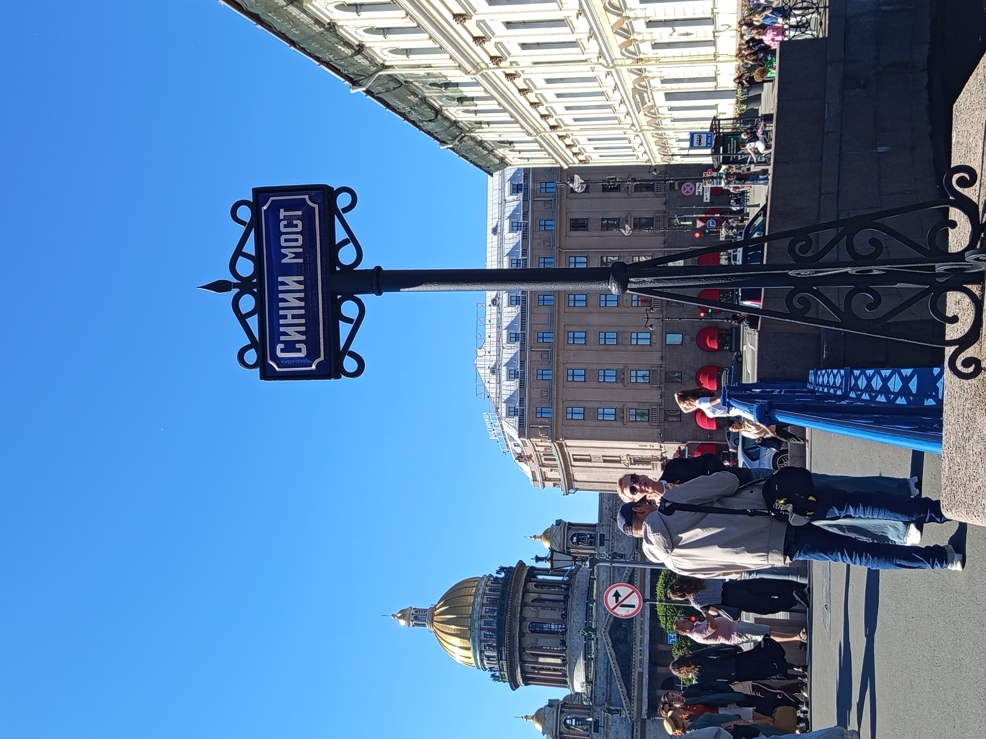 In Saint Petersburg, Russia, on a clear day, a blue street sign with white Cyrillic lettering reading "СИНИИ МОСТ" (Blue Bridge) is prominently displayed on a black metal pole. The sign is ornately decorated with black scrollwork. In the background, the golden dome of St. Isaac's Cathedral is visible, a majestic landmark. Adjacent to the cathedral is a light-brown building with many windows. To the right of the sign, a cream-colored building with a classical facade lines the street. A group of people are gathered near the sign, some engaged in conversation, while others appear to be tourists. A red and white circular sign indicating a turn restriction is also visible. The scene is set on a street with a pedestrian walkway and a body of water, likely a canal or river, with a blue railing in the foreground.