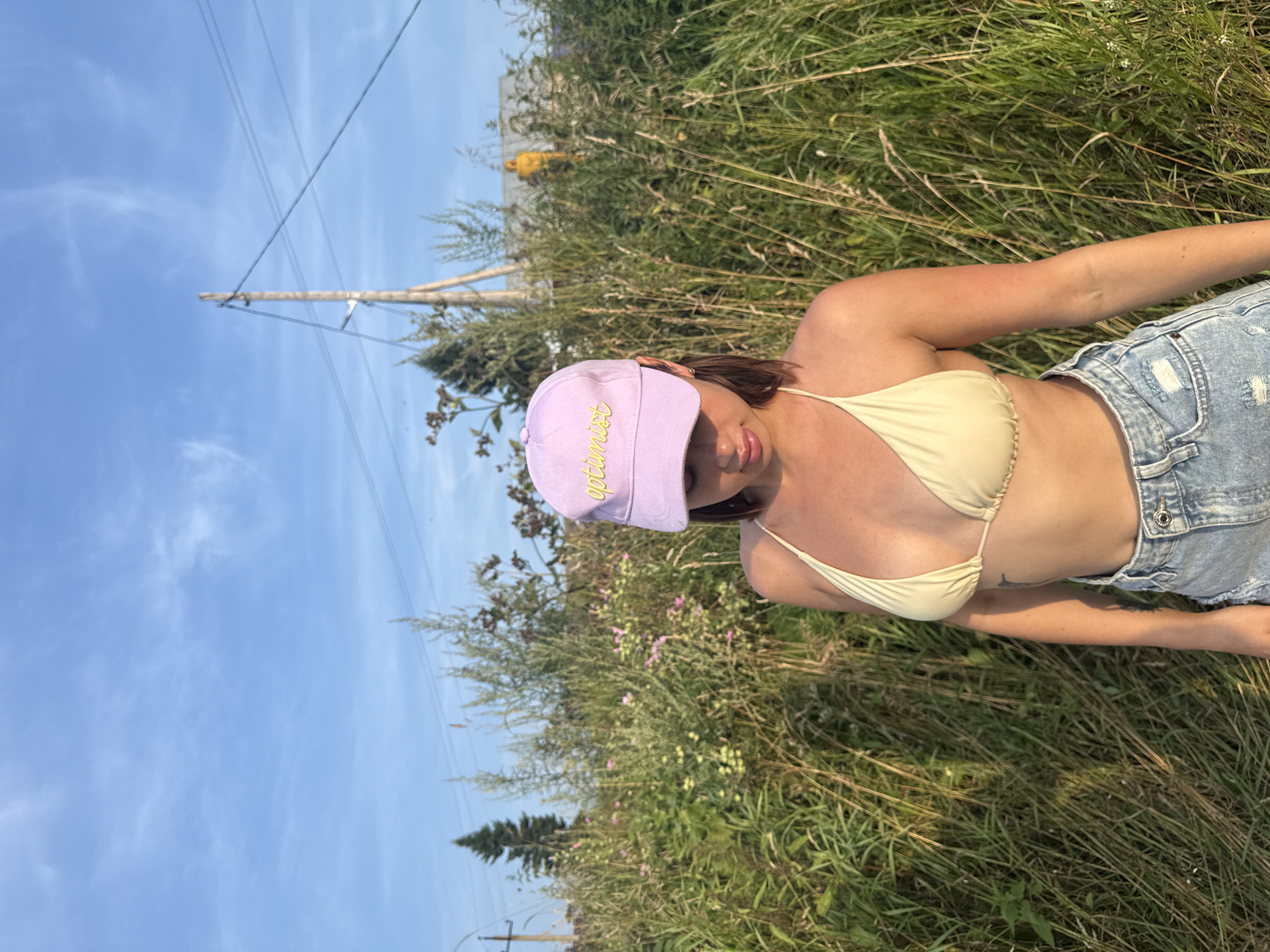 A young woman wearing a light yellow bikini top and denim shorts stands in a field of tall grass and wildflowers. She is also wearing a light purple baseball cap with the word "optimist" embroidered in yellow. The background shows a rural landscape with trees, a wooden utility pole with wires, and buildings in the distance. The sky is blue with wispy clouds. The photograph was taken outdoors during the daytime, likely in Zabor'e, Russia.
