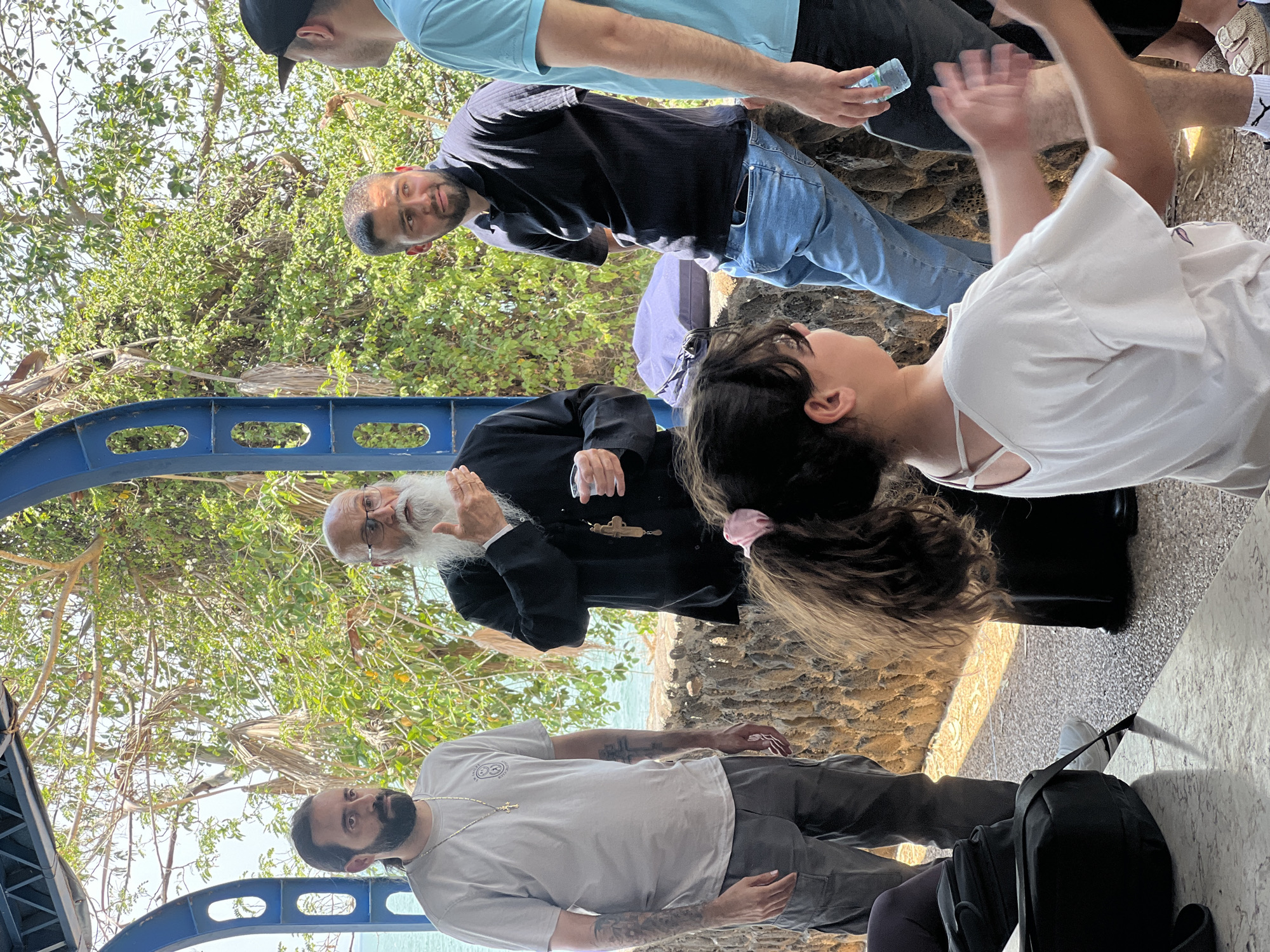 The image shows a group of people standing outdoors near a body of water, likely in the Emek HaYarden Regional Council, Israel. In the foreground, a young girl with long brown hair tied in a ponytail is facing away from the camera, wearing a white t-shirt. Behind her, to the left, a man with dark hair and a beard, wearing a light grey t-shirt and grey cargo pants, is standing. To the right of the girl, an older man with a long white beard and glasses, wearing a black cassock and a wooden cross, appears to be speaking. To the right of the older man, two other men are visible. One, wearing a dark blue polo shirt and blue jeans, is leaning against a stone wall. The other, wearing a light blue t-shirt, is holding a bottle of water. The background features lush green trees and vegetation, with a glimpse of water and sky beyond. The scene appears to be under an open-air structure with blue metal supports.