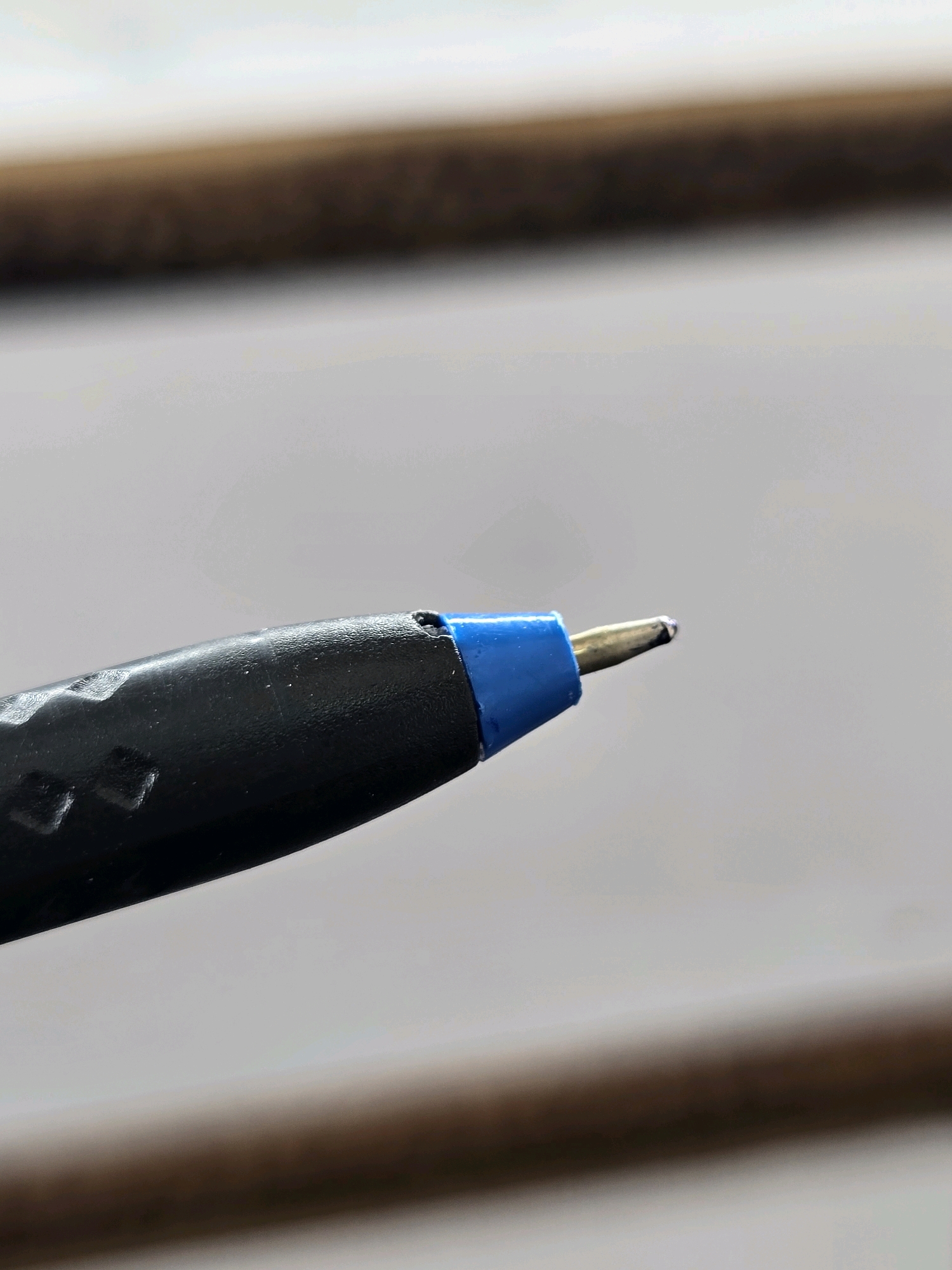 A macro shot shows the tip of a black and blue pen with a metallic nib. The pen has a textured black grip near the tip, with a blue plastic piece separating the grip from the metallic nib. The background is a soft blur of light and dark horizontal stripes, suggesting a textured surface. The image is cropped to focus on the pen's writing end. There is no indication of the city of Thara, India, or any people present.