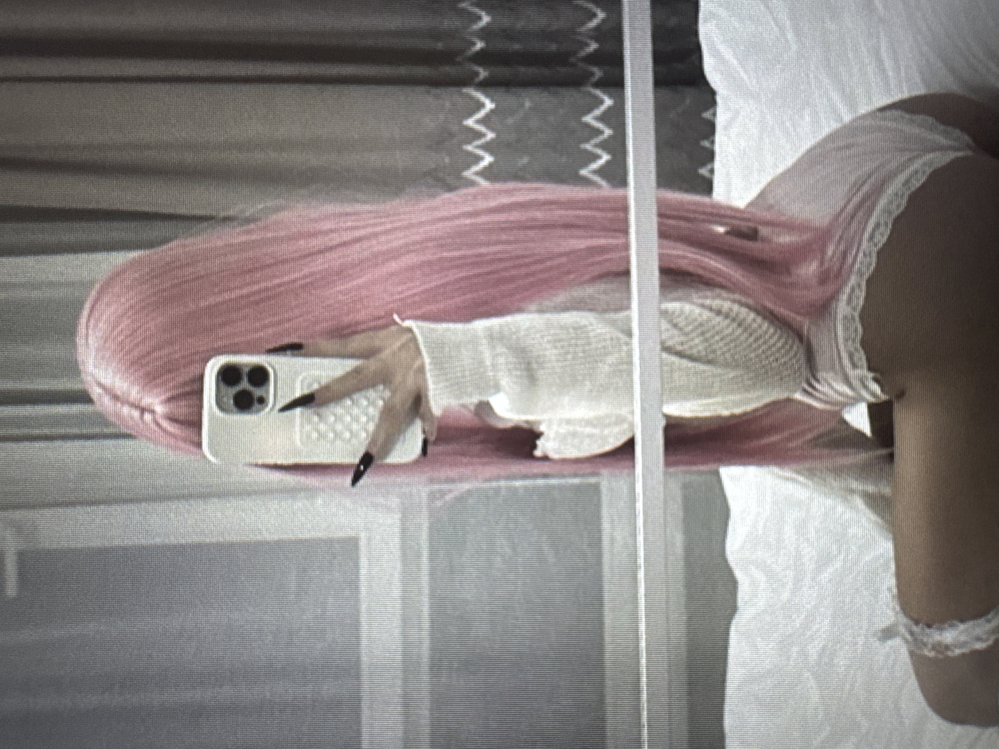 The image displays a reflection selfie of a person with long, flowing pink hair, oriented horizontally across the frame. The individual is wearing a light-colored, possibly white or cream, long-sleeved top with a textured, ribbed or knitted pattern. White, lace-trimmed underwear or shorts are visible on their lower body, with a partial view of their back and upper thigh.
The person's right hand, adorned with long nails featuring black tips, firmly holds a white smartphone. The phone, identifiable by its triple-lens camera system, is encased in a white, textured cover, possibly studded or quilted, and is being used to capture the reflection.
The reflected background features dark, textured surfaces that resemble a wall or curtains, exhibiting faint white zig-zag patterns. A prominent vertical light-colored beam or frame appears to divide the reflection. On the right side, the person is situated on a soft, white surface, likely a bed or sofa. A white, lace-like accessory is visible around their upper thigh or wrist.
The overall aesthetic is characterized by a muted color palette and a slightly grainy quality. No specific time of day, weather conditions, or discernible emotional states are apparent. There is no visible text within the image, and no visual cues directly link the scene to Varna, Bulgaria.