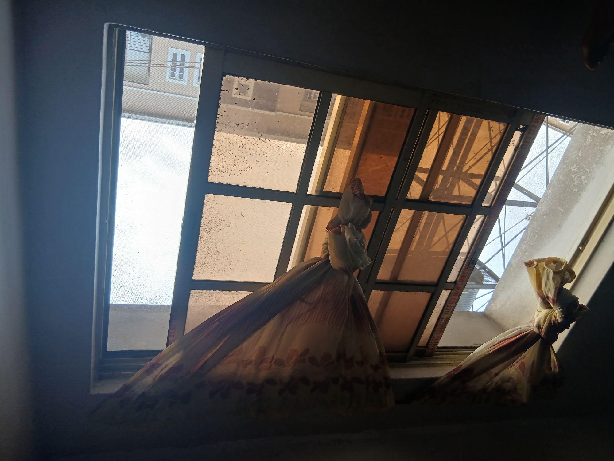 The image shows a window in a room in Lagos, Nigeria. The window is divided into panes, with a view of a building across the street visible through some sections. The window is partly covered by a tied-up fabric, possibly a curtain, which obscures the view through other sections. The light is uneven, with some areas appearing bright and others shadowed. The window frame and glass appear dirty.