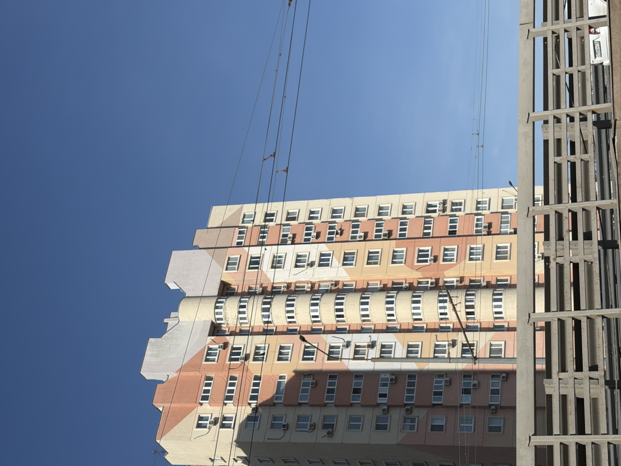 The image depicts a tall building in Kazan, Russia, viewed from a low angle against a clear blue sky. The building is multi-storied with a patterned facade of beige and peach, adorned with numerous windows and air conditioning units. A metal structure, possibly part of scaffolding or another building element, is visible on the right side of the frame. Power lines or cables extend from the top of the building towards the sky.