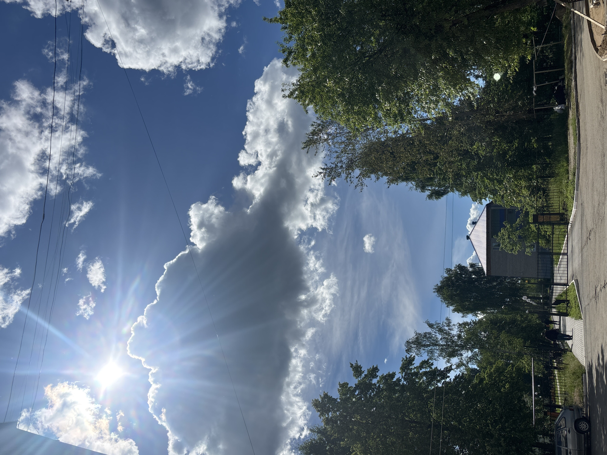 In Chaikovsky, Russia, a bright sun shines through a blue sky filled with fluffy white clouds. Tall trees with green leaves line a paved pathway leading to a metal gate in front of a brick building. A few people are walking on the path, and a silver car is parked nearby. The scene appears to be a residential area on a sunny day.