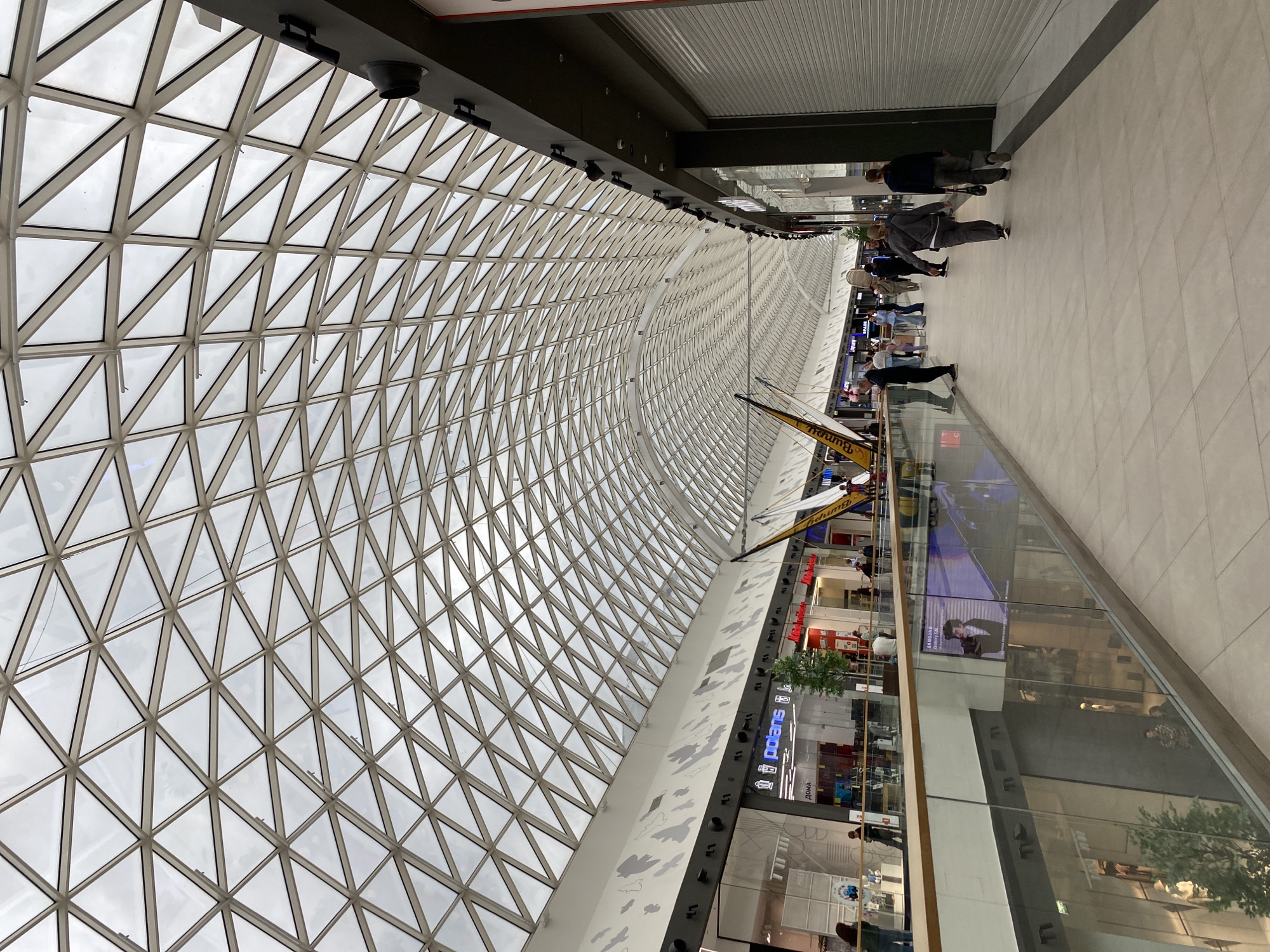 The image shows the interior of the "iMall Esplanada" shopping center in Perm, Russia. The distinctive feature is the arched glass roof constructed of triangular panels. Below the roof, the image shows a long corridor with retail stores on one side and a pathway for pedestrians on the other. People are seen walking through the mall. Some stores visible have signage in Cyrillic. A large advertisement is partially visible, and a piece of machinery or equipment with flags is present within the scene. The overall environment suggests a bright and modern shopping center during daytime hours.