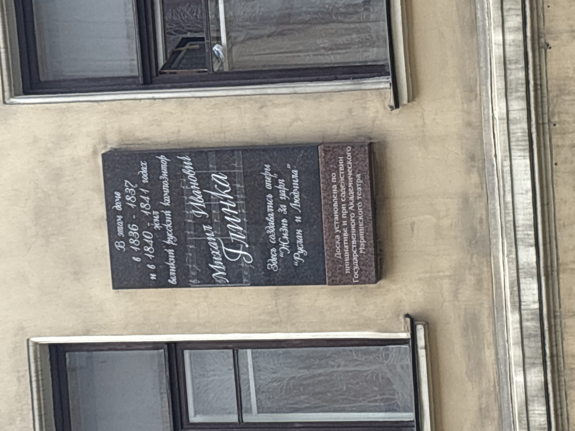 The image shows a commemorative plaque on the facade of a building, likely in Saint Petersburg, Russia. The plaque is dark gray with white lettering and features musical notation. It states that in this house, from 1836-1837 and from 1840-1841, lived Mikhail Ivanovich Glinka, a great Russian composer. It also mentions that here were created the operas "A Life for the Tsar" and "Ruslan and Lyudmila." The bottom part of the plaque indicates that it was installed on the initiative and with the support of the State Academic Mariinsky Theatre. The plaque is mounted on a tan-colored wall, with a dark-framed window visible to the left and right of the plaque. The windows have sheer curtains. The building appears to be a multi-story structure with architectural detailing.