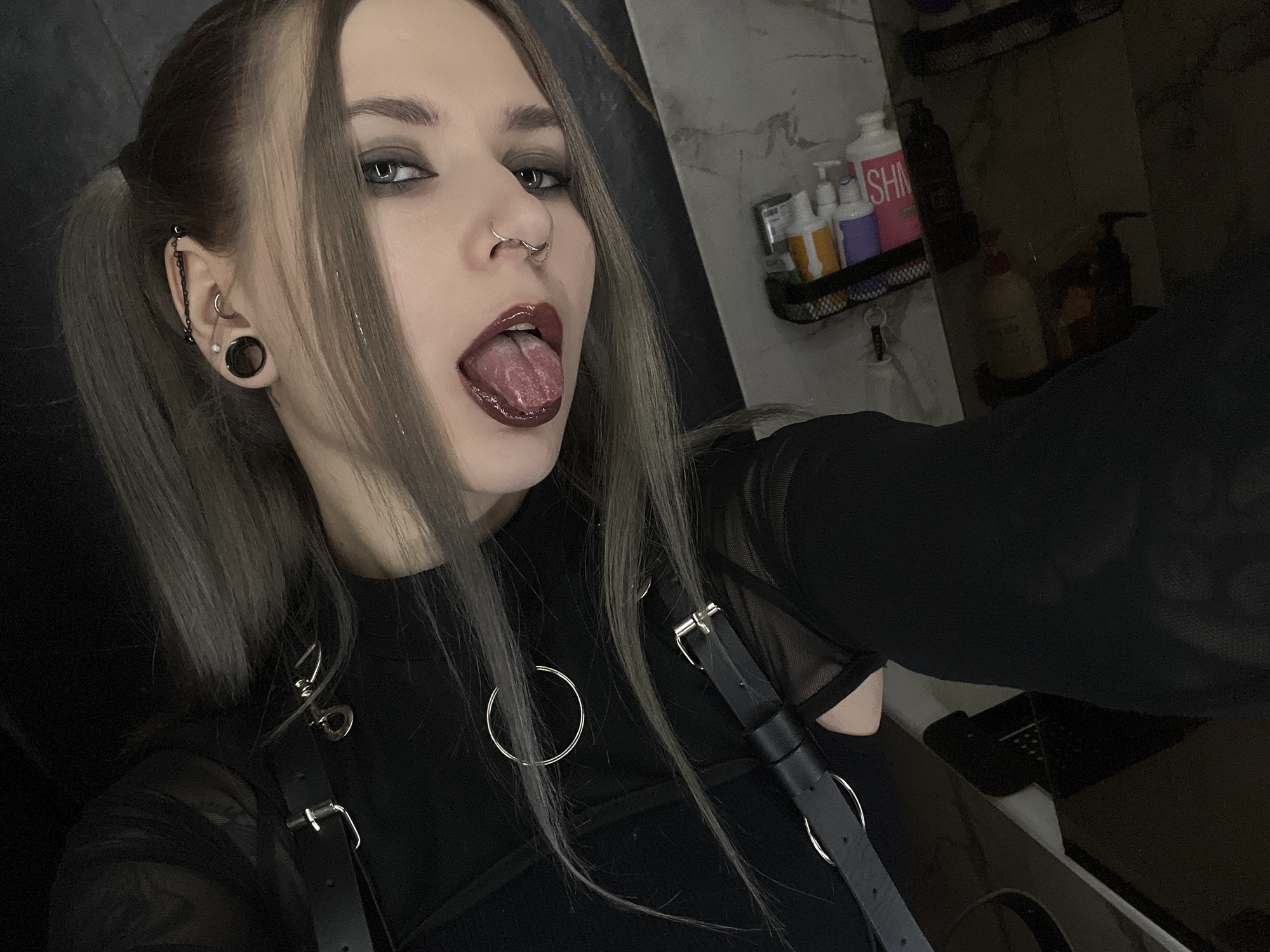 A young female, estimated to be in her late teens or early twenties, is depicted in a selfie, with her tongue partially extended from her mouth. Her hair is medium-length, straight, and appears to be a grey or ash-blonde shade with darker roots, styled in pigtails. Dark eye makeup is applied to her eyelids, and her lips are covered in a dark red or maroon lip gloss. She exhibits multiple facial and ear piercings: a septum ring, two rings on her right nostril, a large black ear tunnel in her left ear, a small hoop in her left upper helix, a small pearl stud below the hoop, and an industrial piercing connected by a black chain in her left ear. She is wearing a black top, which appears to have sheer or mesh sleeves, and a black harness or accessory featuring silver buckles and multiple silver rings, including a large one positioned centrally on her chest. The background reveals an indoor setting, likely a bathroom, characterized by a mirror reflection on the right side of the frame. Within the reflection, a black mesh caddy holds several personal care product bottles, identifiable by their shapes and pump dispensers; one prominent bottle is pink with partial text "SHN" or "SHM", alongside purple, orange, and white bottles. The wall visible in the reflection presents a light-colored, possibly marbleized texture. The scene is dimly lit. The location of this event is in the city of Khimki, Russia.