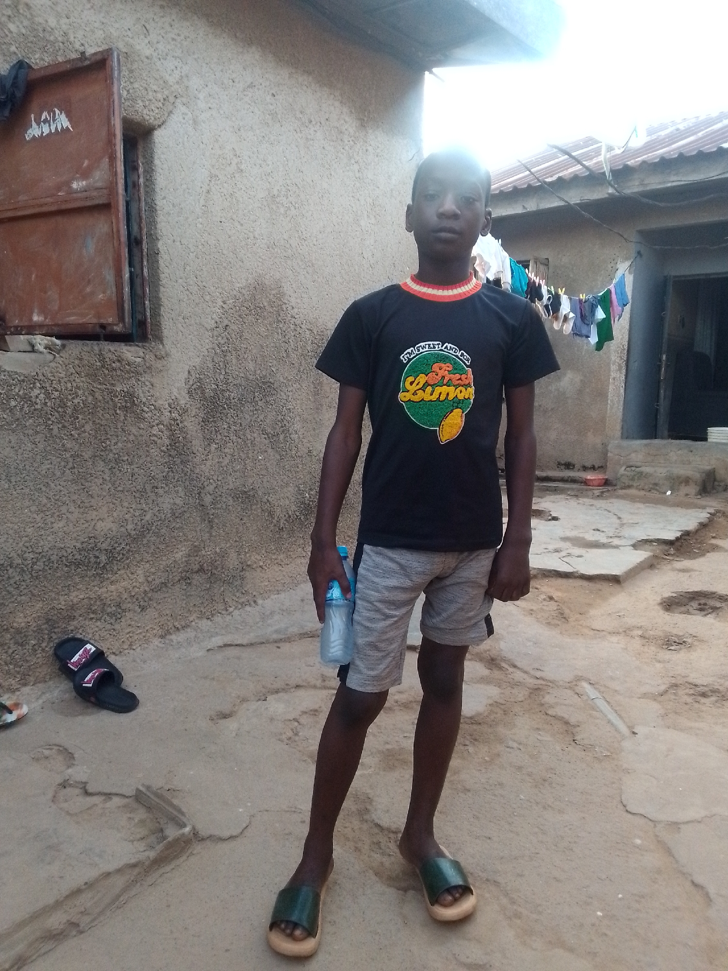 A young boy stands in a courtyard with a concrete floor. He is wearing a black t-shirt with an orange and yellow trim and a graphic of a lemon with the words "I'M SWEET AND FOR FRESH LEMON" on it. He is also wearing grey shorts and dark green sandals on his feet. In his right hand, he holds a clear plastic bottle of water. To his left, a pair of black sandals lies on the ground. Behind him, a rough, stucco-covered wall rises, with a rusty metal shuttered window on the left. Clothes hang on a line in the background. The environment appears to be an outdoor area in a residential setting, possibly in a developing country.