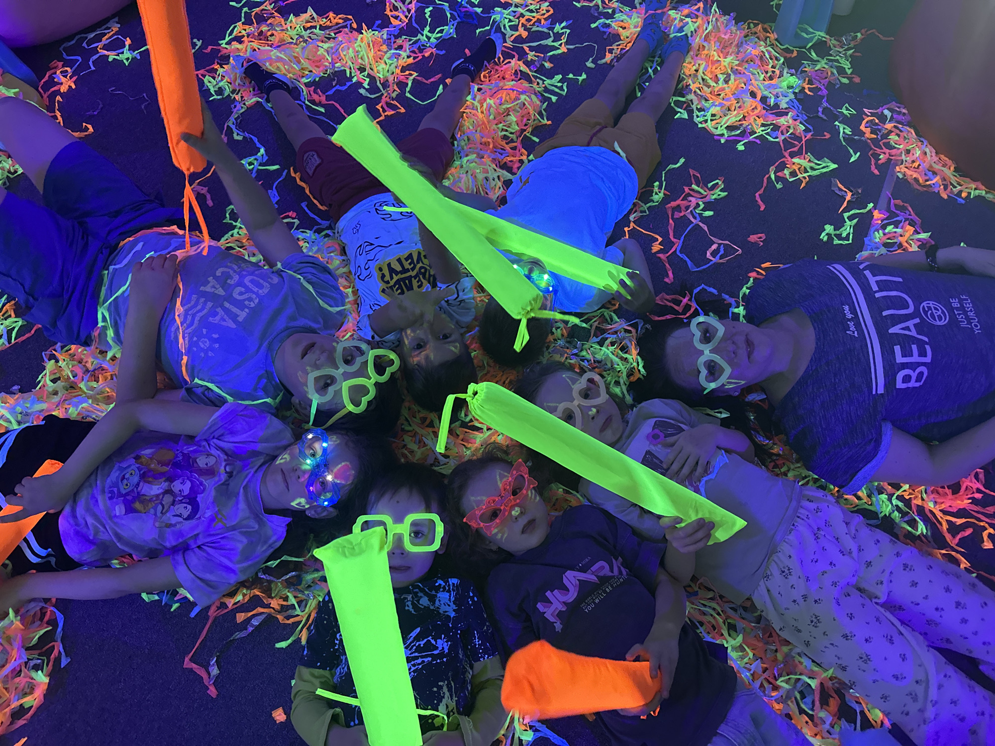**Description:**
The image shows a group of approximately 8 children and one adult lying on their backs on a dark floor covered with brightly colored confetti in a room with UV lighting. They are surrounded by various fluorescent objects, including glow sticks (orange and green), glasses with heart or square frames, and possibly other glowing decorations. The atmosphere suggests a party or celebration setting, likely indoors.
The children are diverse in age and are wearing an assortment of casual clothing, from t-shirts and shorts to pajamas. The one adult is wearing a t-shirt with the words “BEAUT” and “JUST BE YOURSELF” and pajama pants. The scene is likely from a party or fun event due to the glow-in-the-dark elements and party streamers.