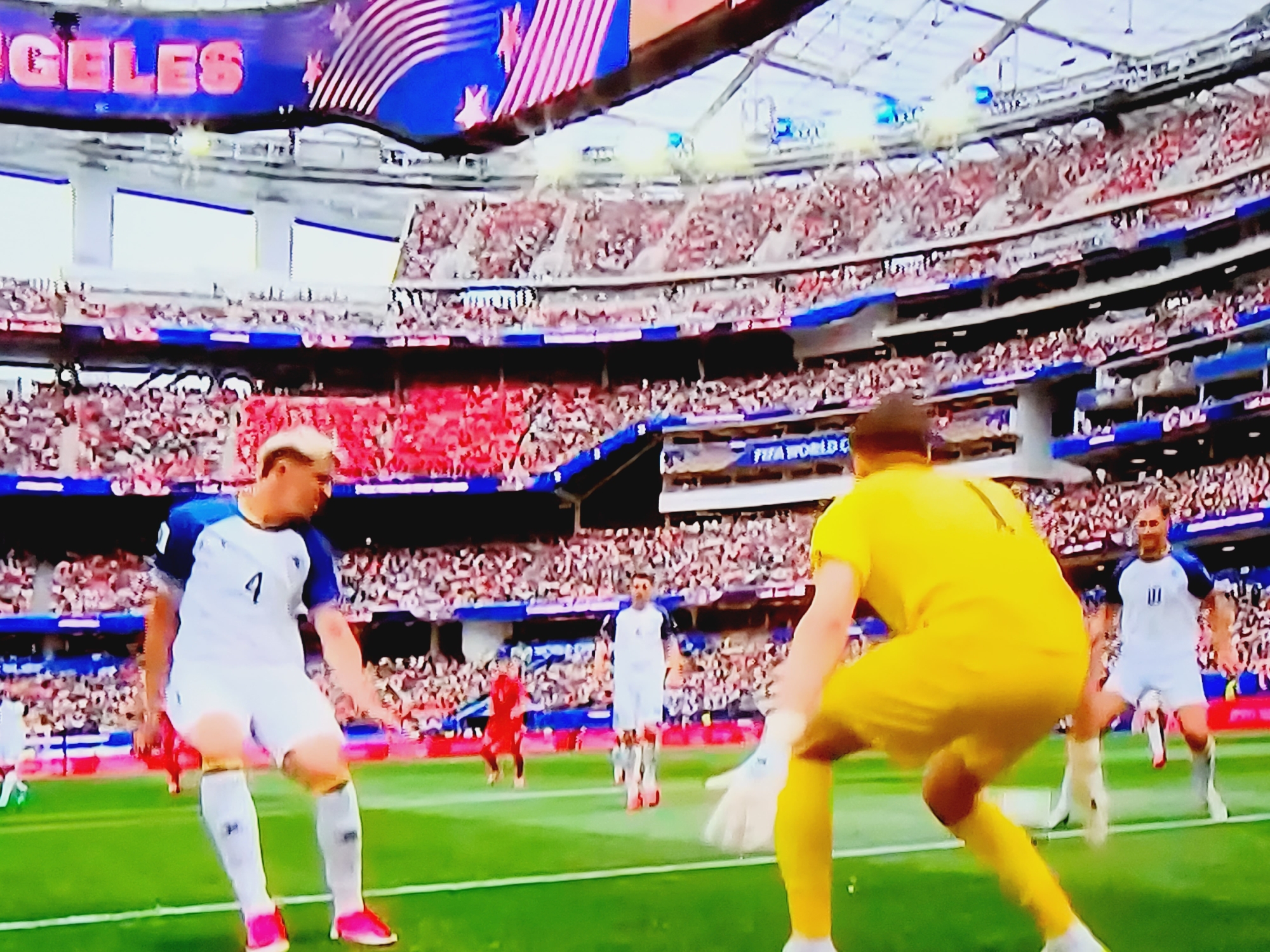 The image depicts a football match in a stadium filled with spectators. The stadium's ceiling features a large banner with the text "LOS ANGELES" in pink lettering, along with an American flag motif. In the foreground, two football players are visible on the green field. One player, wearing a white jersey with blue accents and the number 4, is in motion, leaning forward with his arms out. The other player, in a yellow jersey and pants, appears to be a goalkeeper, facing away from the camera with his hands extended. Behind them, more players in white and red jerseys can be seen, as well as a densely packed crowd of spectators in the stands. The overall scene is a dynamic snapshot of a live sporting event. This image is not located in Baku City, Azerbaijan.