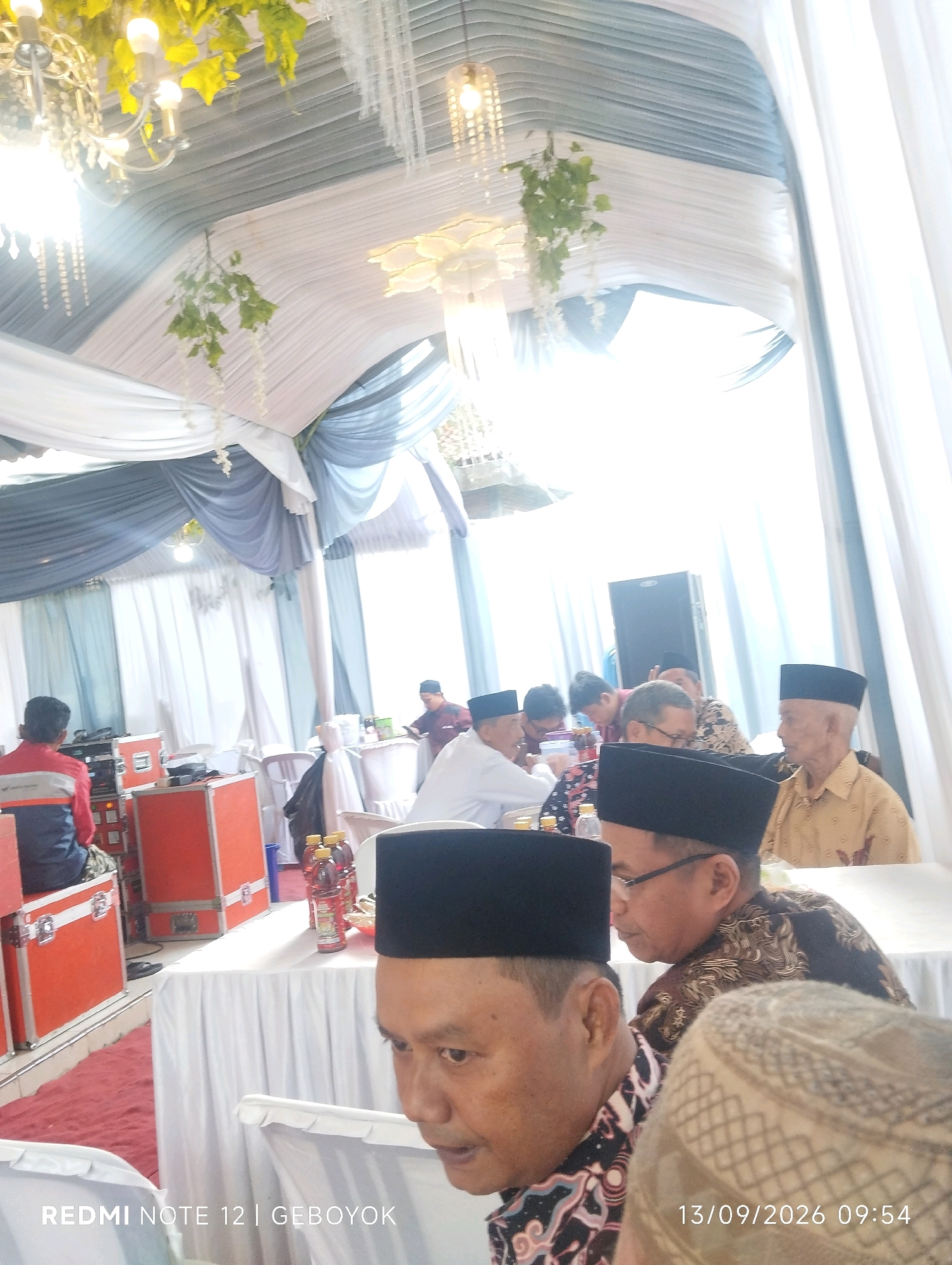 The image captures a gathering, likely a wedding reception, set up under a draped tent in Kuwukan, Indonesia. Several men are seated at tables covered with white cloths, some wearing peci hats. The decor features white and gray fabric draping, chandeliers, and artificial greenery. In the foreground, a man wearing a black peci hat and a patterned shirt is partially visible. To the left, equipment cases are stacked, and a person in a red and blue jacket is seated. Bottles of beverages are placed on a table. The date stamp "13/09/2026 09:54" suggests a future event.