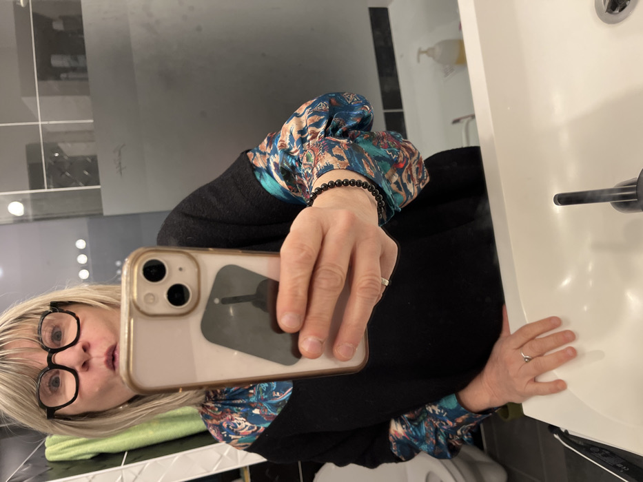 A person takes a selfie in a bathroom mirror. The person has blonde hair, black glasses, and wears a black sweater with patterned blue, green, and brown sleeves, and a black beaded bracelet. The phone case is gold. A white sink with a black faucet is visible in the foreground. The background shows a modern bathroom with a mirror reflecting the surroundings. A white dispenser is mounted on the wall.