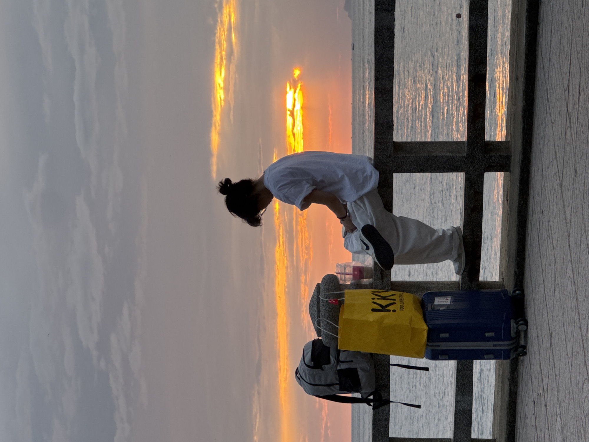 A person sits on a railing overlooking the ocean at sunset in Nha Trang, Vietnam. The sky is filled with dramatic clouds illuminated by the setting sun, creating a warm, orange glow. The person, viewed from behind, is wearing a white t-shirt and light-colored pants, with their hair pulled up in a bun. To their left, a grey backpack, a yellow shopping bag with "KKI 100 LIFESTYLE" printed on it, and a blue rolling suitcase are neatly stacked on the railing. A cup with a straw is also visible near the shopping bag. The ocean stretches out to the horizon, with a faint silhouette of land in the distance. The railing is made of dark stone, and the walkway in the foreground is paved with patterned tiles. The overall scene evokes a sense of peaceful contemplation amidst a vibrant natural display.