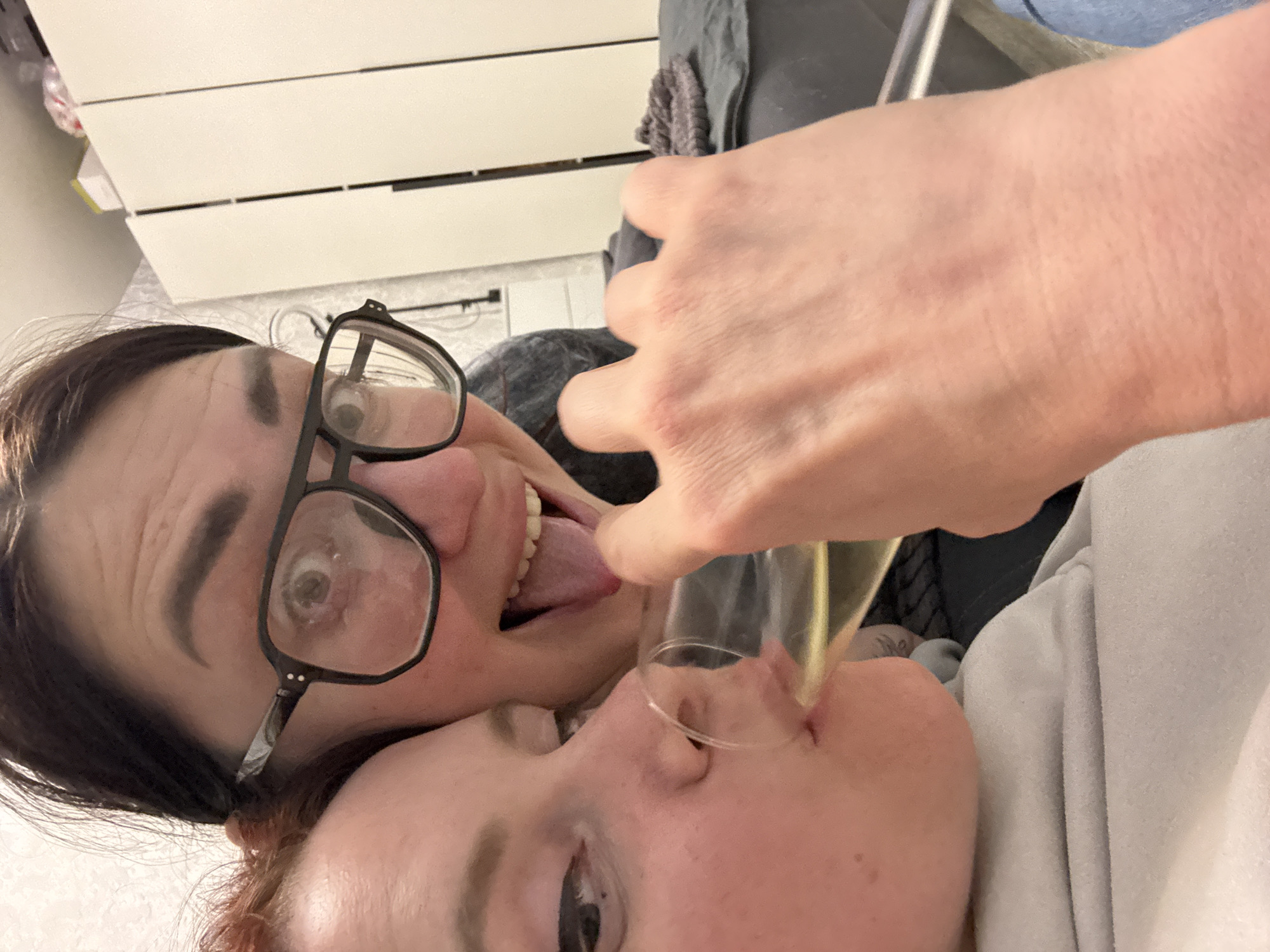 Two people are visible in the image. One person wears glasses and has dark hair. They are sticking their tongue out. The other person is lying down and has red hair. Someone is holding a glass up to their nose, as though to pour something inside. The location appears to be indoors, near a white cabinet.