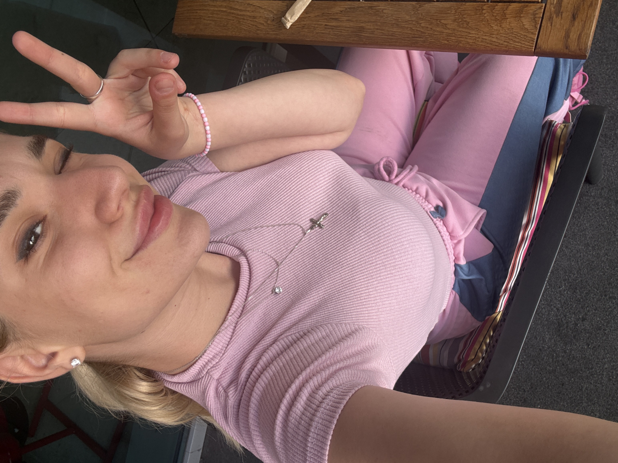 A young woman is depicted in a close-up selfie, seated outdoors in what appears to be a chair with a dark frame, possibly on a balcony or patio in Sofia, Bulgaria. A wooden surface, resembling a table or railing, is visible in the upper portion of the frame, above her head and arm. The bright natural light suggests it is daytime with clear, sunny weather.
She has light skin, blonde hair visible around her ear, and dark eyebrows. Her left eye is playfully closed in a wink or a wide smile, and her lips are slightly parted, conveying a cheerful and relaxed emotion. She is wearing a light pink, ribbed short-sleeved top. Her lower body is clad in pink loungewear, possibly sweatpants, with visible drawstrings, and a blue panel or garment is also noticeable on her lap. A striped cushion, featuring red, yellow, black, and white stripes, is tucked into the chair behind her.
For accessories, she wears a silver ring on her right hand's ring finger and a pink and white beaded bracelet on her right wrist. Her right hand is raised, forming a peace or victory sign with her index and middle fingers. Around her neck, she has a thin silver chain necklace with two pendants: a small, sparkling solitaire and a larger, silver bird-like or abstract shape. A small, clear stud earring adorns her left earlobe. There is no visible text in the image.