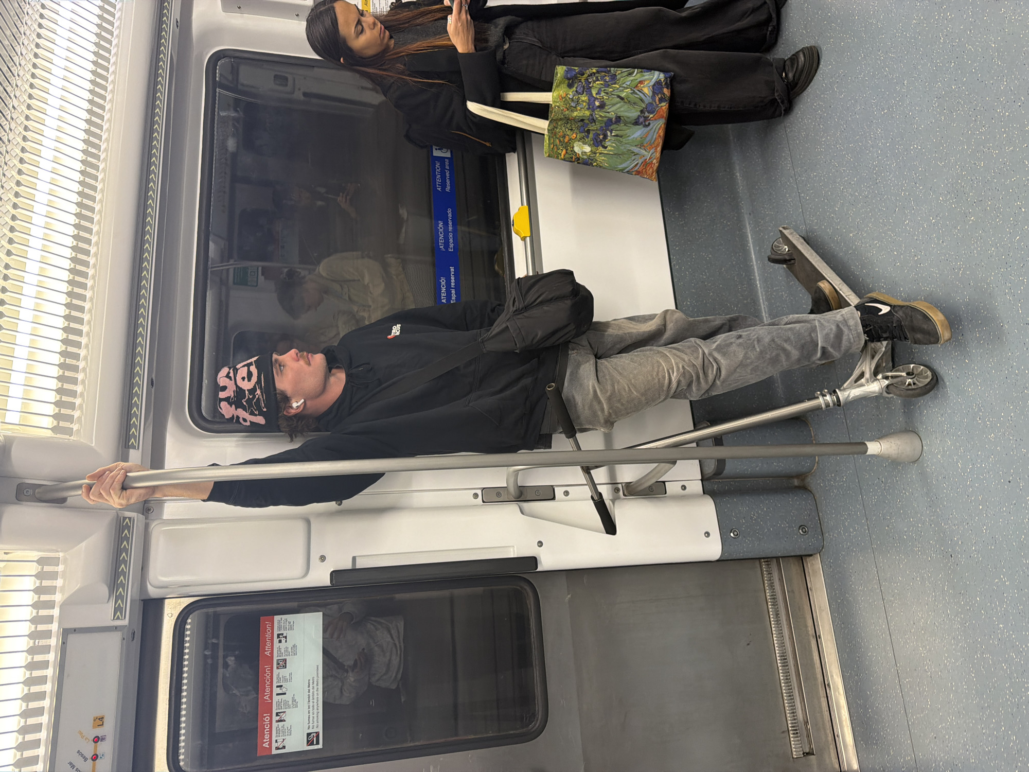 The interior of a subway car shows a man lying down along a side bench, his head near a vertical pole he's gripping with his hand. He's wearing a black hoodie and light gray pants, with a patterned hat and what appears to be an earbud in his ear. Next to his feet is a scooter. A woman stands near him, dressed in black and holding a bag that seems to have a floral print. Signage in Spanish, Catalan, and English related to reserved seating can be observed above the man. Some of the signage includes Besòs Mar, Besòs, La Pau, and L4.