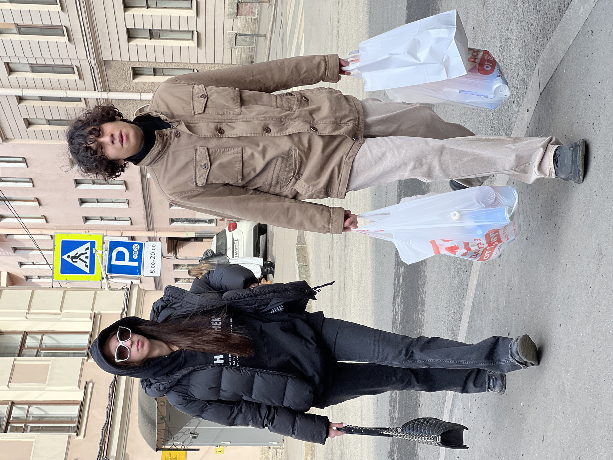 Two individuals walk on a paved sidewalk in Saint Petersburg, Russia, against a backdrop of historic multi-story buildings and street signs. The scene is illuminated by daylight, indicating clear visibility.
The individual on the left is a female with long dark hair, wearing a black hooded puffer jacket over a black hooded sweatshirt with partial white text, dark wash jeans, and dark chunky sneakers. She wears white-framed sunglasses and carries a black quilted shoulder bag with a silver chain strap in her left hand. Her gaze is directed forward.
The individual on the right, walking slightly ahead, is a male with curly dark hair, wearing a tan/khaki long-sleeved field jacket over a black turtleneck, light beige pants, and dark chunky sneakers. He carries a transparent plastic shopping bag containing items, including what appear to be bottles or cans, in his left hand, and a white paper shopping bag in his right hand. His head is turned slightly to his right.
In the background, a white Mercedes-Benz sedan with Russian license plate B461PX 178 is parked on the street. Overhead, a blue square pedestrian crossing sign with a white icon and a blue square parking sign indicating "P" with "8.00-20.00" in Cyrillic are mounted on a pole. The buildings behind the subjects feature windows with light-colored frames and varying shades of pink and beige facades.
