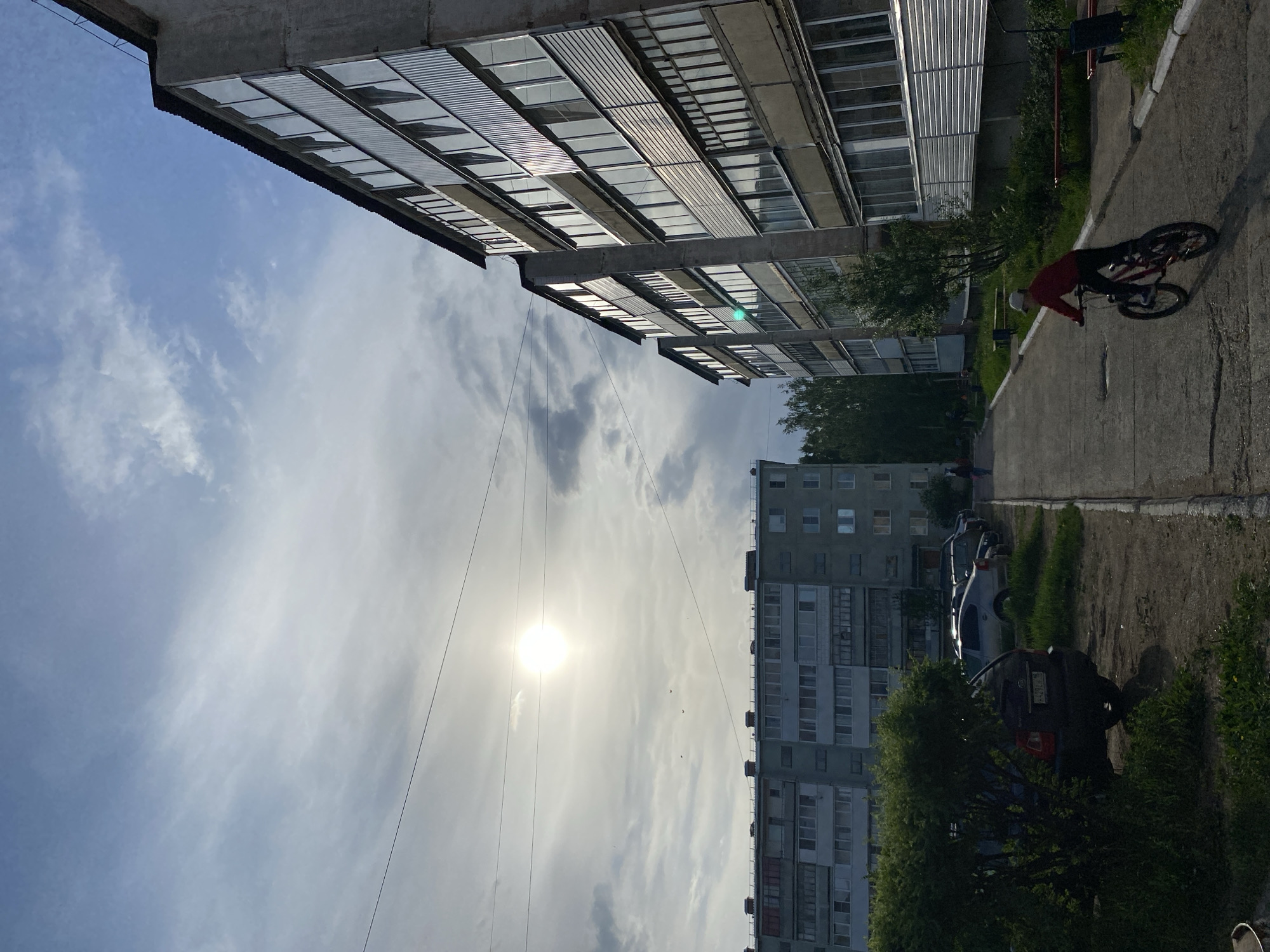 An exterior shot taken from a low angle showcases buildings, a street, and a sky filled with clouds. The photograph appears to have been taken with the camera rotated counter-clockwise. Buildings line the scene's periphery: one building is located in the top, and another is oriented along the central portion of the photograph. Parked vehicles are present in the bottom section of the image. On the right side, an individual in red is riding a bicycle, positioned on a pavement with grass growing along the edge. Power lines are visible against the sky. A bright sun is visible through the clouds. The location is the city of Noviy, Russia.