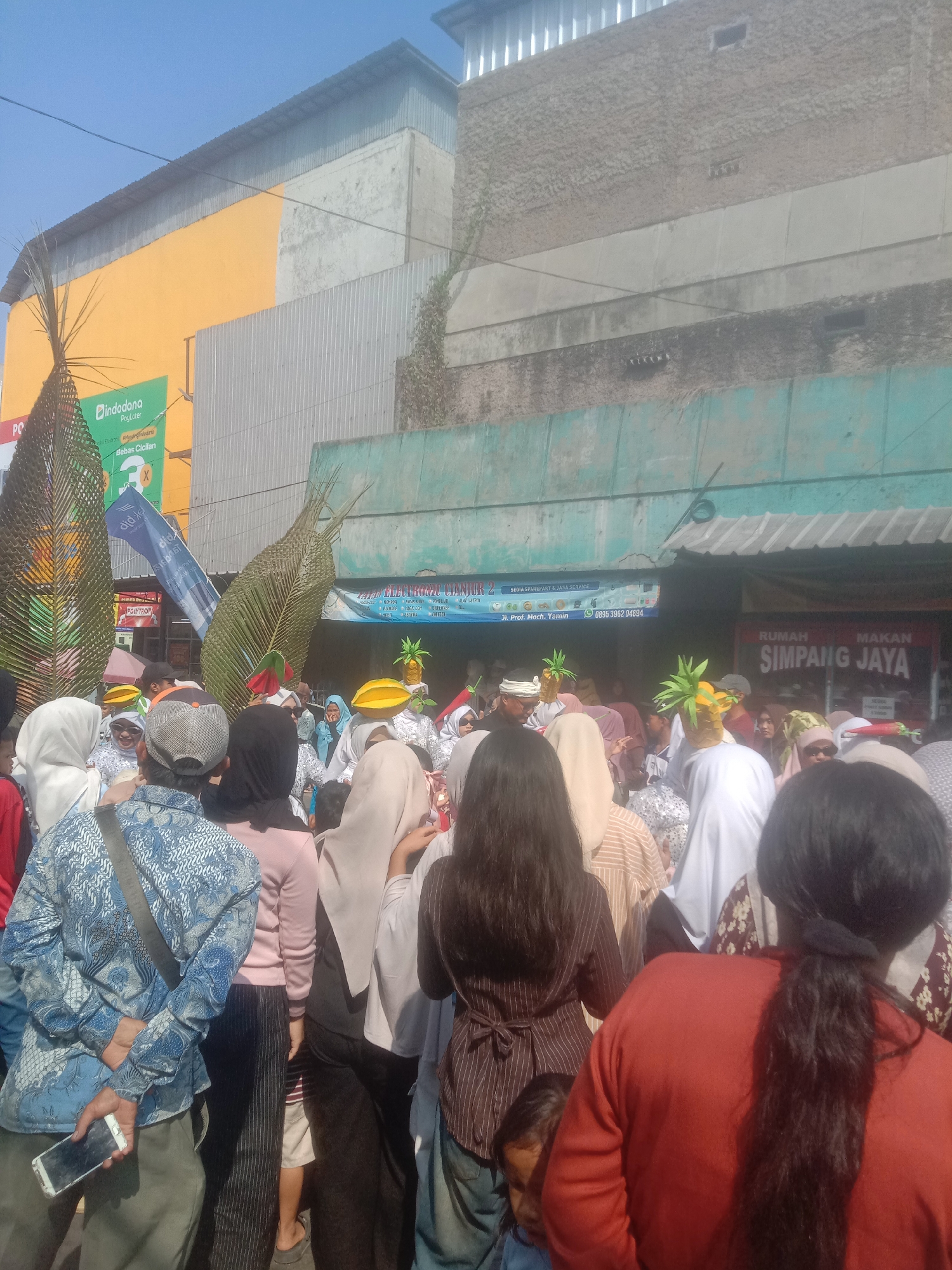 In Cianjur, Indonesia, a crowd of people is gathered outdoors, possibly for a parade or festival. Large, stylized leaf-like decorations made of woven material stand on either side of the street. Some individuals in the crowd wear distinctive headdresses shaped like fruit, such as pineapples and mangoes. Banners are visible in the background, one advertising "Indodana PayLater" and another for "RUMAH MAKAN SIMPANG JAYA." The buildings in the background are a mix of colors, including yellow and teal, and appear to be commercial establishments. The sky is clear and blue, suggesting a sunny day. The people are dressed in casual clothing, and many appear to be looking towards the front of the gathering, indicating some event is taking place. One person in the foreground is holding a smartphone.