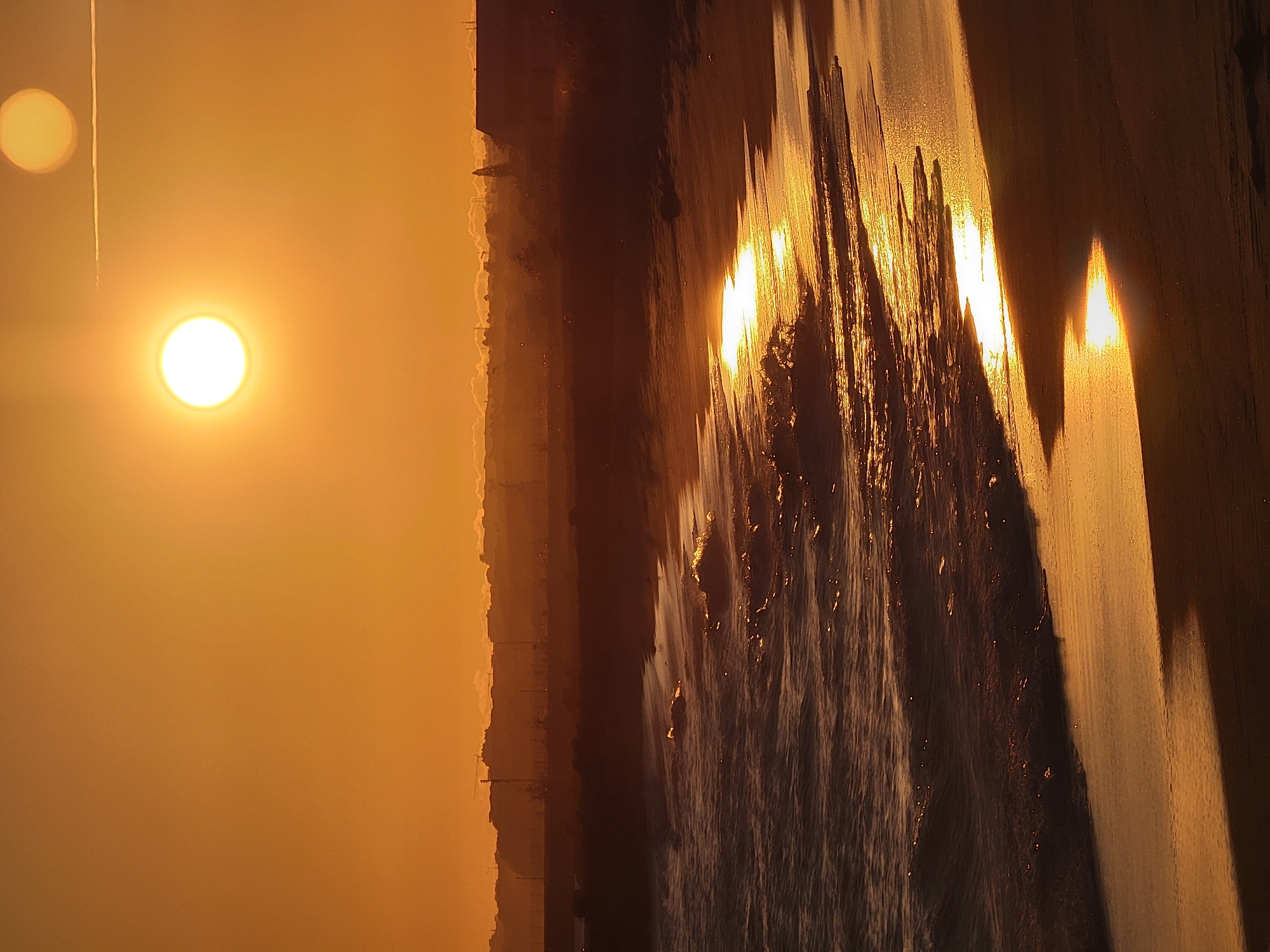 The image depicts a sunset over the Atlantic Ocean at Quarteira, Portugal. The sun, a bright orb, is setting on the horizon, casting a warm, golden glow across the scene. The sky is a vibrant orange and yellow, with the sun creating a reflection on the water and the wet sand. Gentle waves are rolling onto the shore, with the sunlight catching the crests of the waves and the wet sand. In the background, a silhouette of the Quarteira coastline is visible, with buildings and some industrial structures, possibly cranes from the port, dotting the landscape. The overall atmosphere is serene and tranquil, showcasing the natural beauty of the coastal landscape at dusk. There are no people or animals visible in the image.