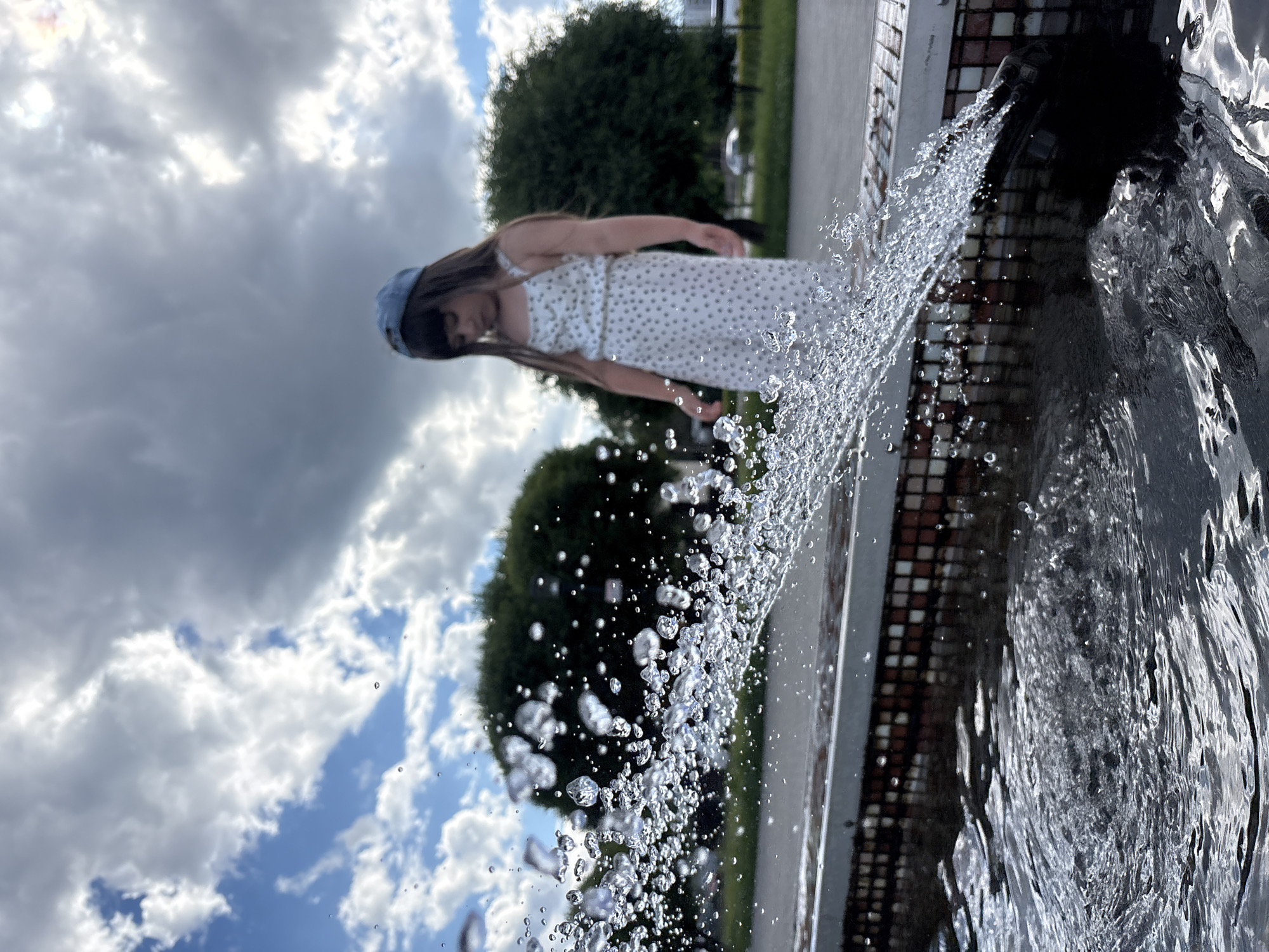 In Chaikovsky, Russia, a young girl wearing a white polka-dot dress and a blue baseball cap stands by a fountain. She looks down towards the water, which is splashing vigorously in the foreground. The fountain's edge is adorned with small, colorful tiles. Lush green trees and a cloudy sky form the background of the scene.
