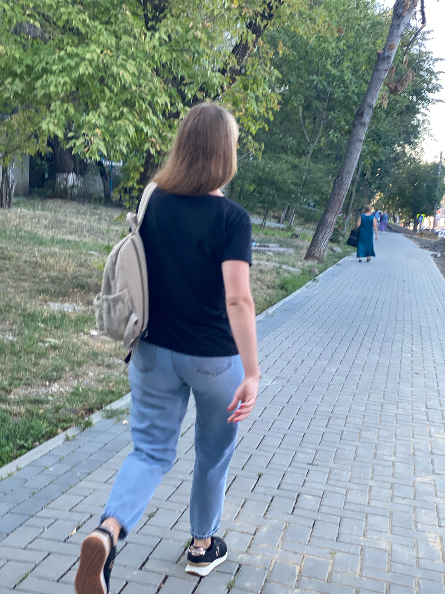 A woman with long brown hair, wearing a black t-shirt and light blue jeans, walks away from the camera down a paved sidewalk. She carries a light-colored backpack over her right shoulder. Her left foot is forward, and she is wearing black and white sneakers. The sidewalk is lined with green grass and trees. In the distance, another woman in a blue dress and a person in a lighter blue outfit are walking away from the camera. The scene appears to be a park or a tree-lined street. The weather appears to be overcast. The overall environment suggests an urban or suburban setting in Chișinău, Moldova.