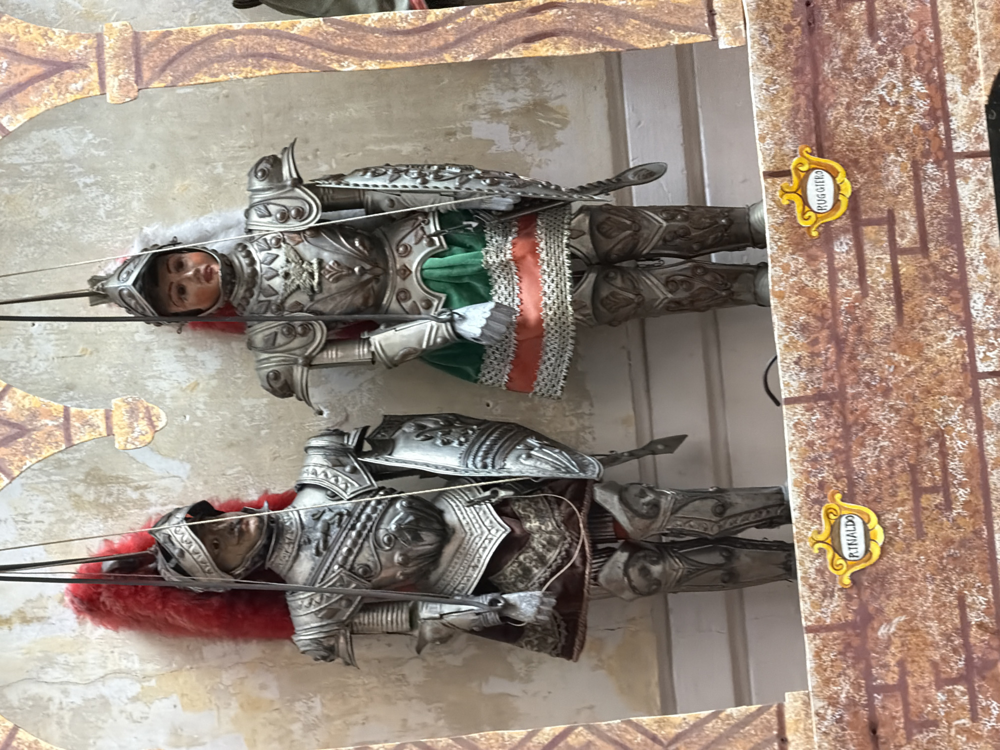 The image shows two marionettes dressed as knights, standing in front of a wall with arched decorations. The marionette on the left is wearing a silver suit of armor, a helmet with a red plume, and a red skirt under its armor. It holds a shield in its left hand. The marionette on the right is also wearing a silver suit of armor, a helmet, and a green and orange skirt. It holds a shield in its left hand and a sword in its right. Below the marionettes, there is a brick-like structure with two decorative signs. The sign on the left reads "RINALDO" and the sign on the right reads "RUGGIERO." The background wall is textured and appears aged, with some areas of peeling paint. The marionettes are suspended by wires, indicating they are part of a puppet show. This scene is located at the Museo delle Marionette in Palermo, Italy, a museum dedicated to the traditional Sicilian puppet theater, known as Opera dei Pupi.