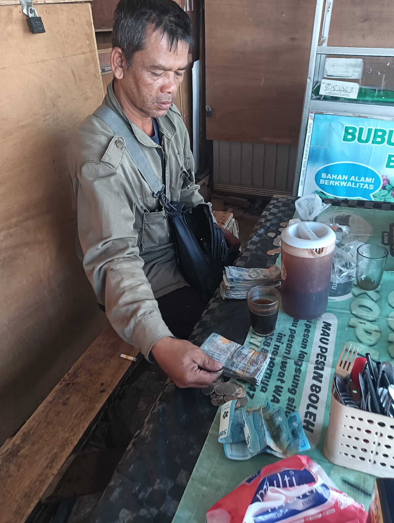 A middle-aged man with dark hair, a mustache, and a tanned complexion is sitting at a table with a checkered tablecloth in what appears to be a small shop or eatery in Gandasari, Indonesia. He is wearing a tan, button-down shirt with pockets and a gray strap across his chest. He is looking down and counting money with his right hand, which is holding a few banknotes and coins. A lit cigarette is visible on the table in front of him. On the table, there is also a glass of dark liquid, possibly coffee, a large plastic container of the same liquid, two small glasses, a stack of Indonesian Rupiah banknotes, loose coins, and a white basket filled with cutlery. In the background, there is a wooden wall and a display case with some signage. One sign is blue and white with text that reads "BUBULK" and "BAHAN ALAMI BERKUALITAS". Another sign has green text and reads "MAU PESAN BOLEH". The scene suggests a casual transaction or a break from work.