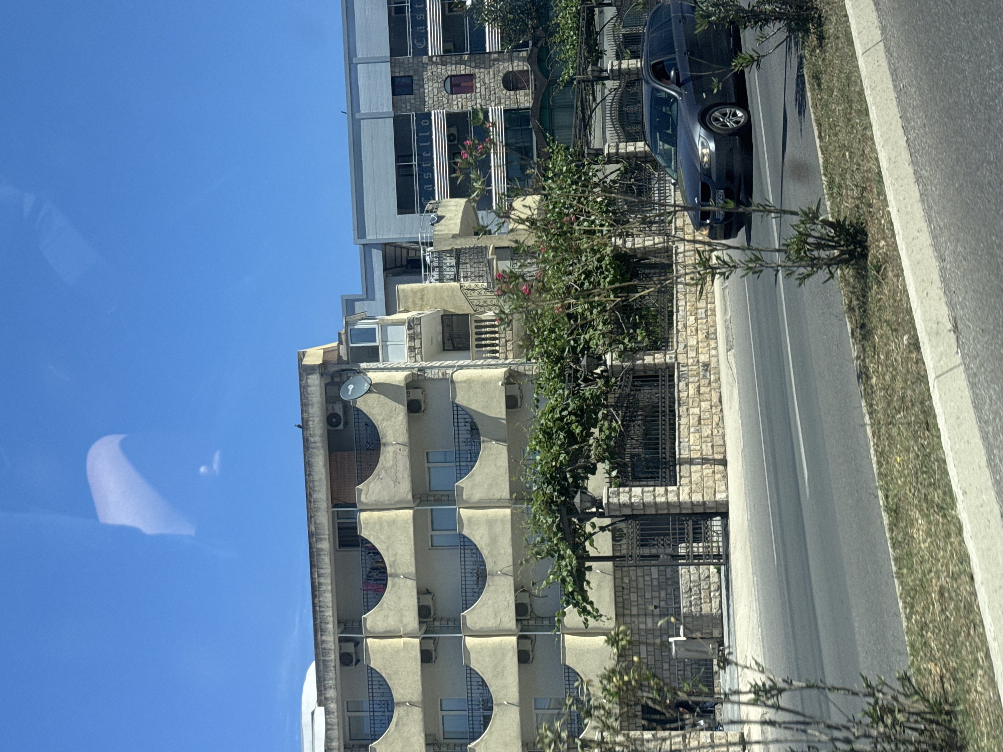 A daytime shot shows apartment buildings in Bar, Montenegro. The buildings on the left are light beige with semi-circular balcony alcoves, each equipped with individual air conditioning units. Adjacent is a taller, grey building with rectangular windows. A black car is parked along the curb, next to a sidewalk and a road. A wrought iron fence runs along the front of the beige building. The sky is blue.