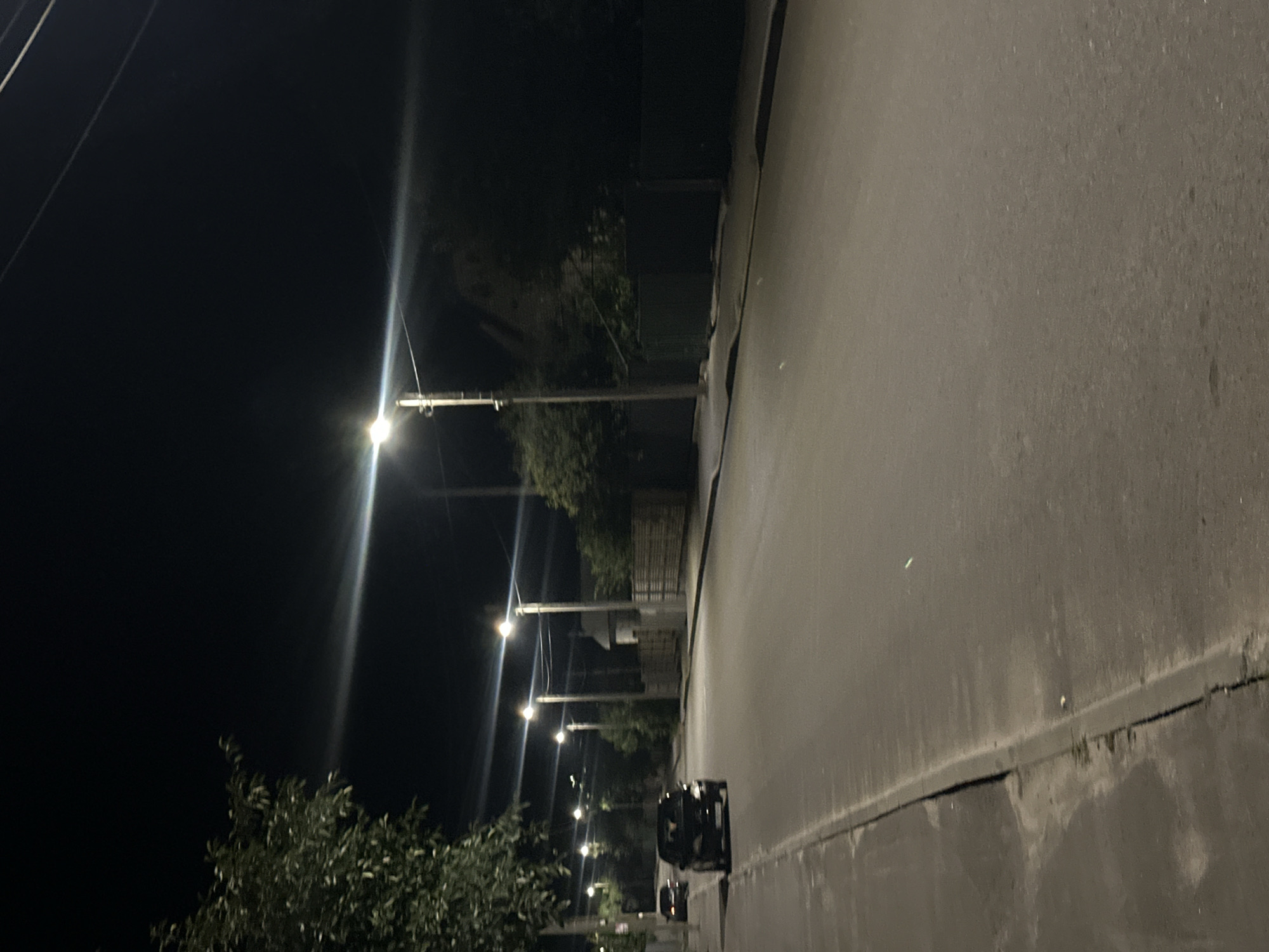 The image depicts a street scene at night in Khmilnyk, Ukraine. Several streetlights illuminate the asphalt road, casting long shadows. On the left side of the road, a sidewalk is visible, with a parked dark-colored car and another car in the distance. Trees and vegetation line the side of the street, with a wooden fence visible further down. The sky is dark, indicating nighttime.