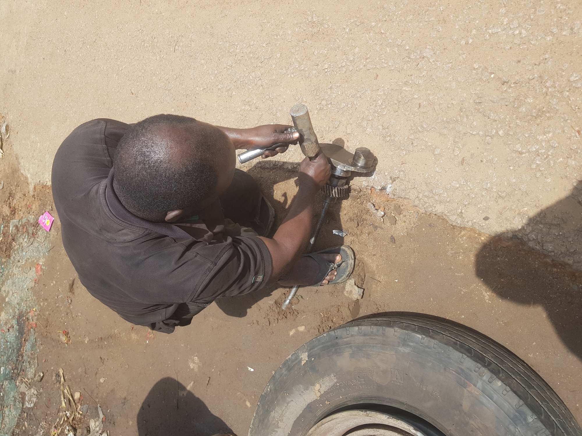 In the city of Tukur Tukur, Nigeria, an individual is seen kneeling on a dusty, rough ground, with a worn tire lying to their left. The person, dressed in a black t-shirt and dark pants, is focused on a metal object in their hands, appearing to be working on a mechanical component with a cylindrical shaft and a gear-like structure. The environment suggests an outdoor workshop or roadside repair area, with debris scattered on the ground, including what looks like broken glass and dry vegetation. The lighting indicates it is daytime, with strong shadows cast on the ground.
