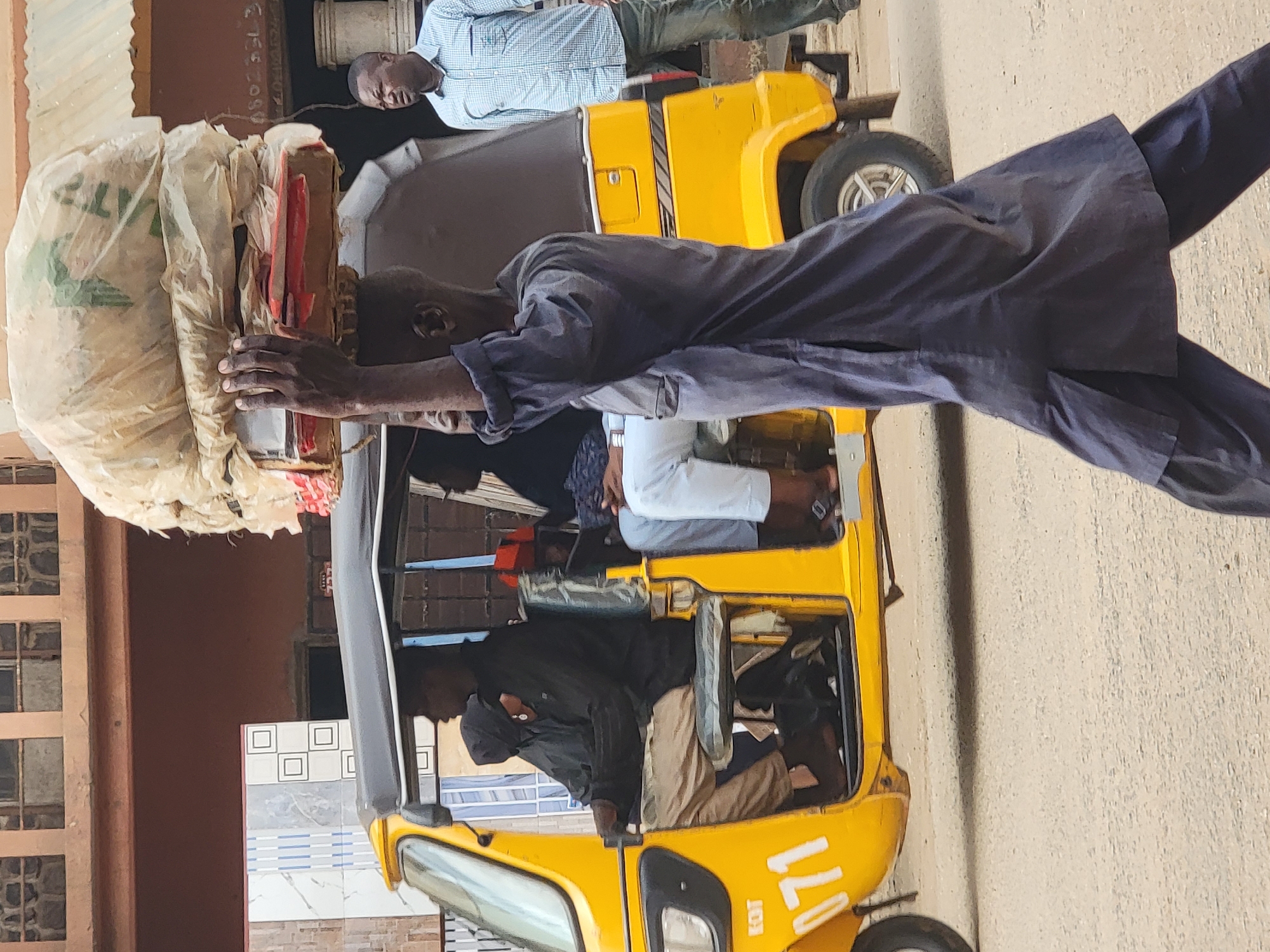 This image captures a busy street scene in Maiburiji, Nigeria, during daylight hours under clear weather conditions. The primary subject is a man, dressed in a dark blue long-sleeved top and trousers, who is skillfully balancing a large, bulky bundle wrapped in light-colored plastic or fabric on his head. He holds the bundle with both hands for stability and appears to be in motion, possibly transporting goods. Within the bundle, a red object is partially visible.
To the man's left, a yellow three-wheeled auto-rickshaw, commonly known as a "keke Napep" in Nigeria, is either parked or moving slowly. Inside the vehicle, several individuals are visible. One person, possibly the driver, wears a dark jacket, while another passenger is seen wearing white pants. Towards the rear of the vehicle, another man in a light blue checkered shirt and dark trousers stands, observing or waiting.
The background features various elements of an urban environment. On the far left, a brown building with multiple window panes is visible. Further back, there are walls adorned with decorative tiles, featuring white squares and blue striped patterns. On the side of the yellow keke Napep, the numbers "71" are distinctly visible. The ground appears to be a light-colored, possibly dusty, surface. The scene portrays a typical moment of daily life and commerce in a Nigerian town.