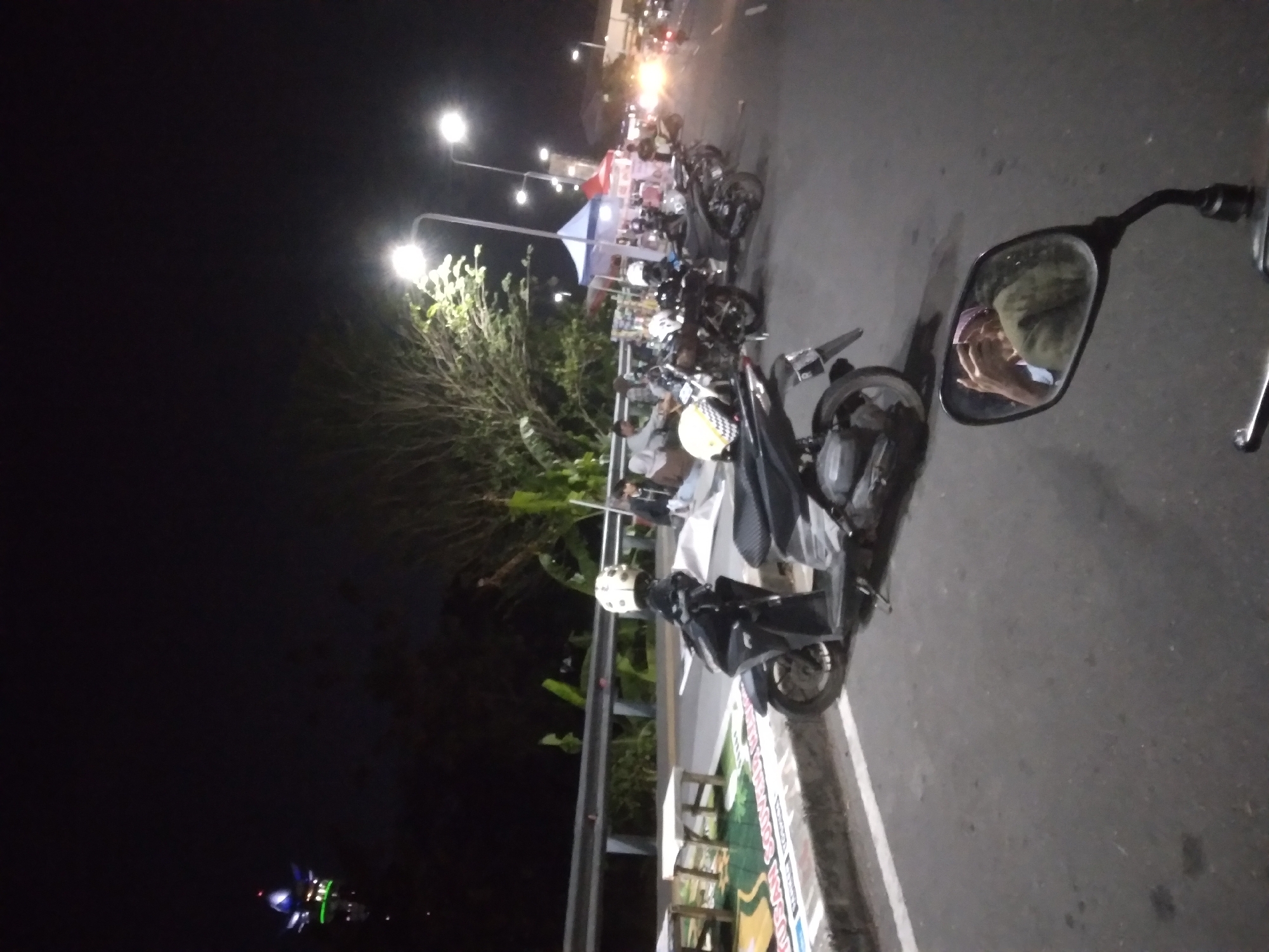The image depicts a nighttime scene on a street in Purwokerto, Indonesia. In the foreground, a motorcycle with a white helmet on its seat is parked on the asphalt road. To the left, there are two white tables and a sign with Indonesian text. Further down the road, several other motorcycles are parked, and people are gathered around small tents, suggesting a market or food stalls. Streetlights illuminate the area, and a distant tower with blue and green lights is visible. The scene conveys a lively atmosphere typical of a night market or a roadside gathering.