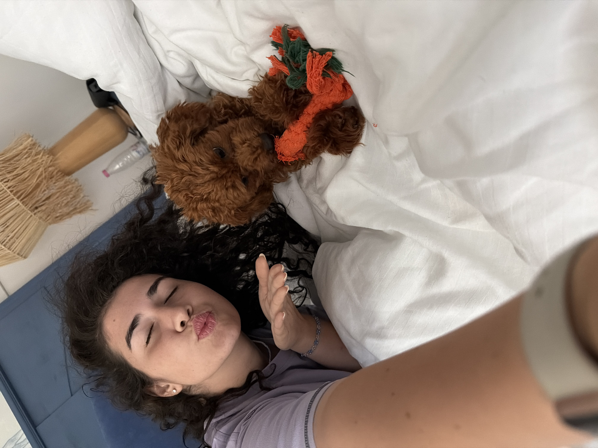 In a bedroom, a young woman with curly dark hair is lying in bed, blowing a kiss towards the camera. She is wearing a light purple t-shirt and a beaded bracelet on her left wrist. Next to her, a small, fluffy red dog is lying on the white duvet, chewing on an orange and green rope toy. The room is bright, with natural light filtering in. The background shows a headboard, a lampshade, and a water bottle on a bedside table. The overall scene suggests a casual and intimate moment between the woman and her pet. The specific location of the bedroom in the city of Banya, Bulgaria, cannot be determined from the visual information provided in the image.