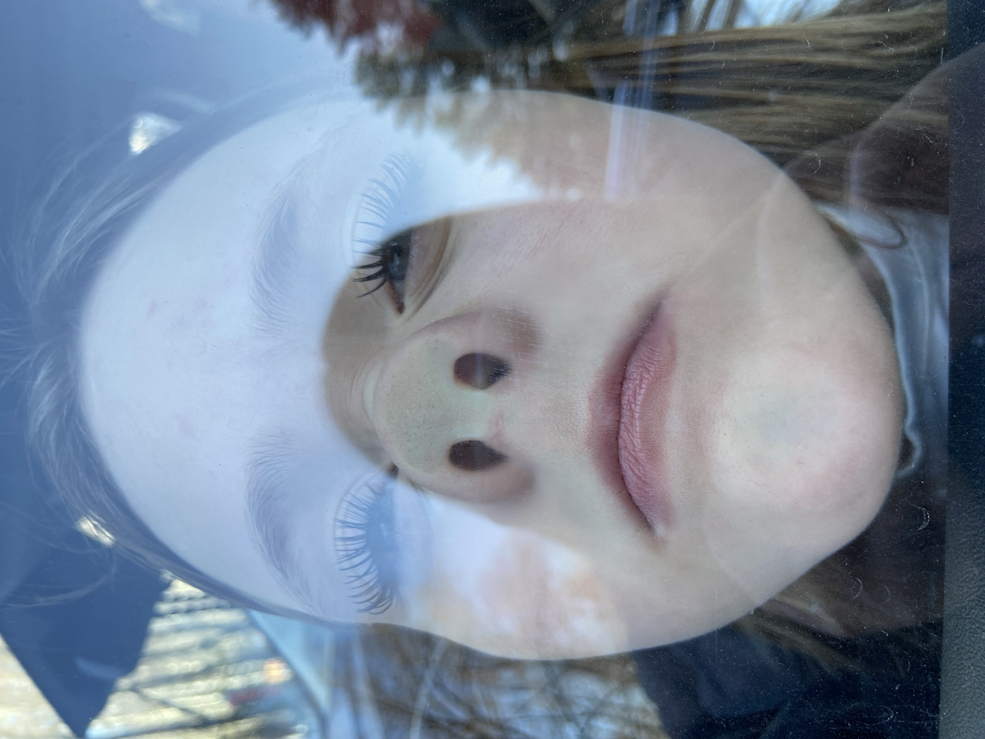 A close-up, inverted reflection of a young person's face, likely a child or young adolescent, is captured on a translucent, reflective surface, possibly a window. The face dominates the frame, displaying fair skin, open eyes with prominent dark eyelashes, a nose, and slightly parted lips. The expression appears neutral or contemplative.
The reflective surface itself shows minor smudges and faint scratches. Through the translucent reflection, a blurred outdoor background is visible, consisting of bright, diffuse areas suggesting sky or strong daylight, and darker, indistinct shapes that resemble trees or foliage. Darker, solid areas framing parts of the face, particularly on the right and bottom, suggest an interior setting from which the reflection is being cast, possibly a car window frame. The overall lighting indicates it is daytime. No specific activity is taking place beyond the presence of the reflection, and no visible text is present. While the location context is Chaikovsky, Russia, no specific visual cues within the image confirm this.