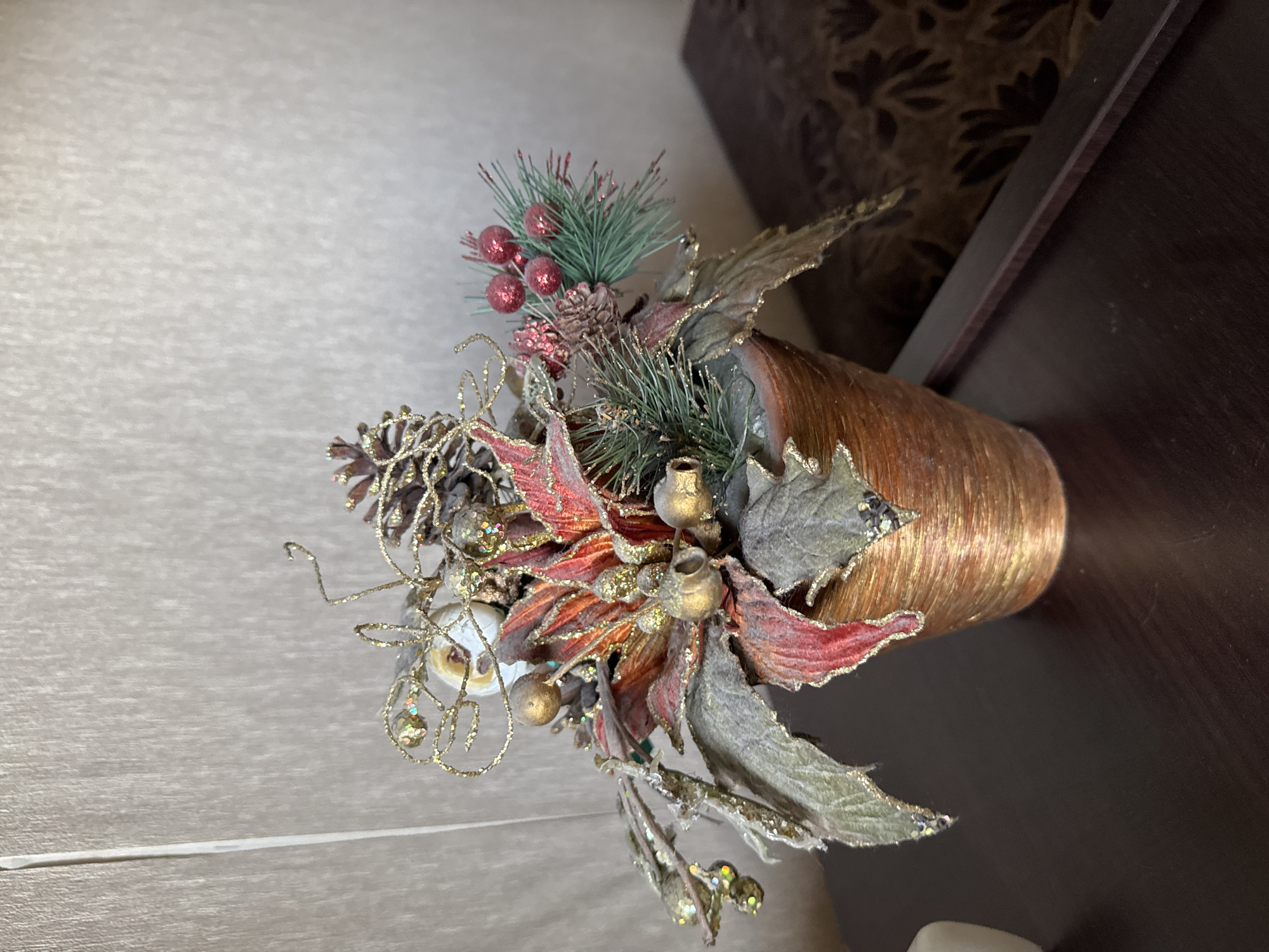 A holiday floral arrangement is centered in the frame, resting on a dark wooden surface. The arrangement features artificial poinsettia leaves in red and green, accented with gold glitter and decorative elements like pinecones, gold beads, and small red berries. The container holding the arrangement is a textured, copper-colored pot. The background consists of a textured, light-colored wall with a vertical white stripe. The scene is indoors.