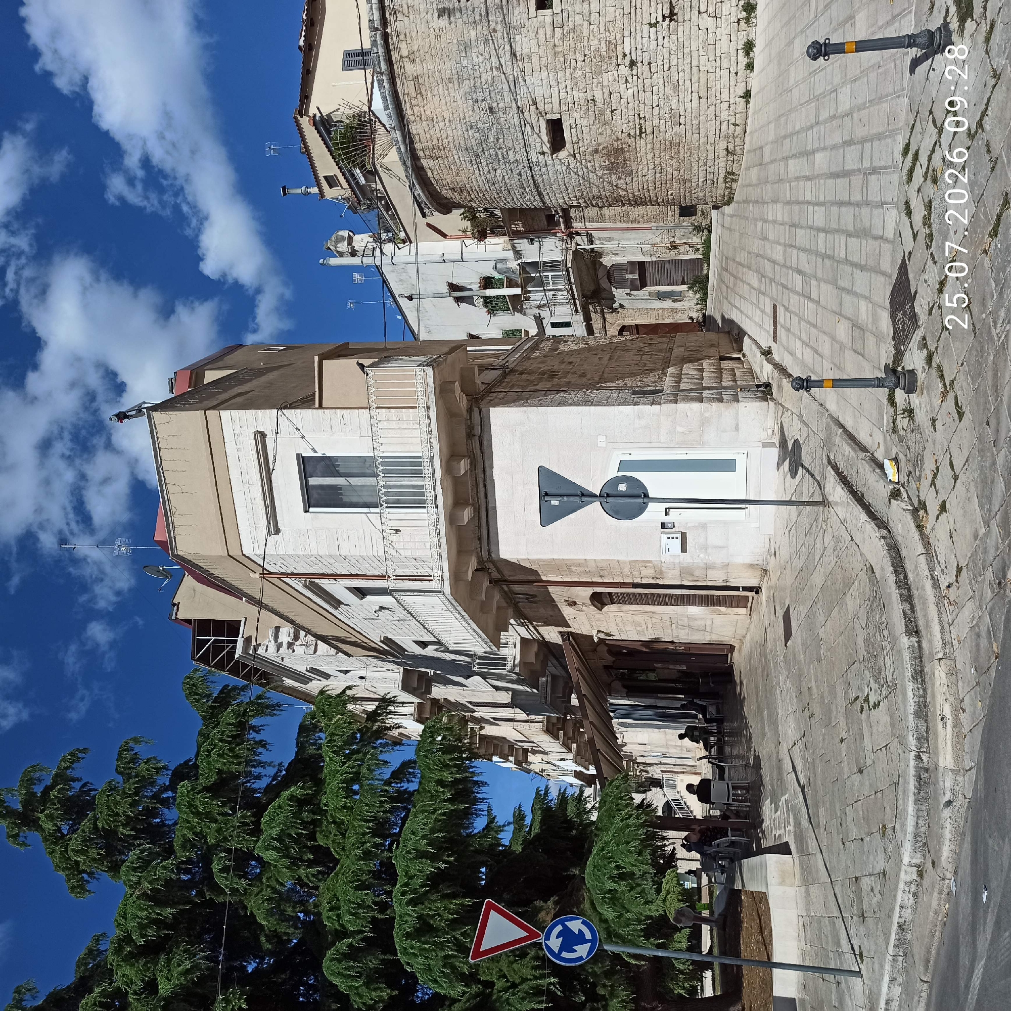 In Ruvo di Puglia, Italy, on July 25, 2026, at 09:28, a view captures an intersection with a large conifer tree on the left, casting a shadow across the paved sidewalk. A white and red triangular yield sign is mounted above a blue circular roundabout sign on a pole. Adjacent to the tree, a two-story building with a light beige facade features multiple windows, some with balconies, and shops or cafes on the ground floor. Several people are seated at tables outside one of these establishments.
The street curves around the corner, leading to a wider area paved with light gray stone tiles. On the right side of the street, a curved wall constructed of rough-hewn light-colored stones forms a prominent feature. Buildings with similar stone construction and balconies are visible further down the street. The sky is blue with scattered white clouds, and the overall scene suggests a sunny day in a European town.