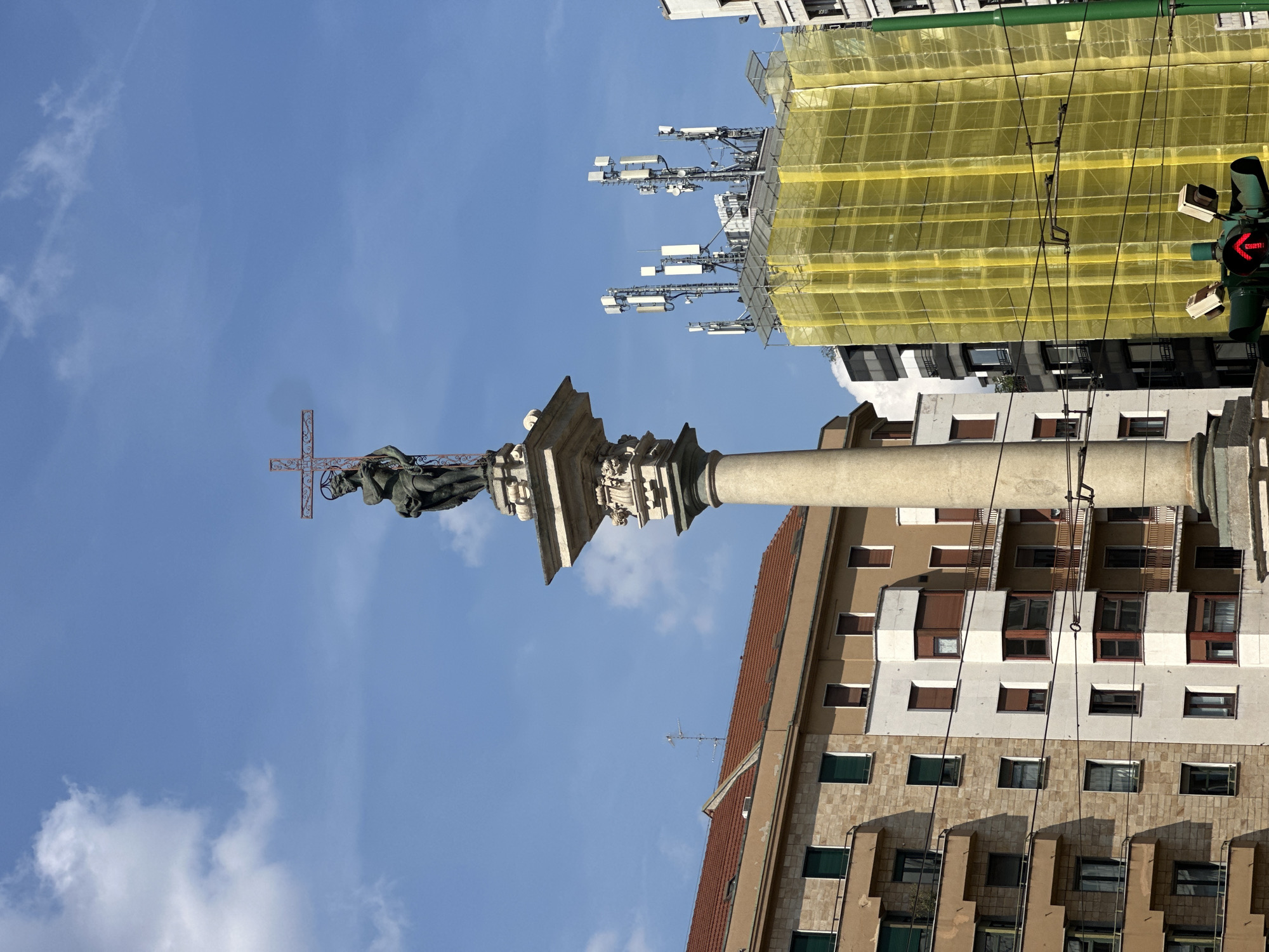The image displays a tall column topped with a statue of Saint John Nepomuk in Piazza San Nazaro in Brolo, Milan, Italy. The central subject is a weathered stone column, likely of Doric or Tuscan order, rising from a decorated pedestal. Surmounting the column is a dark-colored statue of a male figure, presumed to be Saint John Nepomuk, identifiable by the distinctive halo of five stars around his head and the crucifix held in his right arm.
To the left of the column, multi-story residential buildings line the background. These structures feature facades in light brown and cream hues, some with stone cladding and others with smooth render. They exhibit multiple rectangular windows fitted with dark green external shutters and balconies with iron railings. The rooftops are covered with reddish-brown tiles, and a television antenna is visible on one roof.
To the right of the column, a modern building is almost entirely enveloped in bright yellow construction netting, indicative of ongoing renovation or construction work. Perched atop this yellow-covered building is a prominent installation of multiple cellular antennas and telecommunication equipment, consisting of various white rectangular and cylindrical panels mounted on poles. Further to the right, a white building with brown window frames is partially visible.
In the lower right foreground, a green traffic light is visible, displaying a red arrow pointing upwards, signaling a stop for straight-ahead traffic. Dark overhead wires, likely for trams or trolleybuses, traverse the scene horizontally. The entire scene is bathed in bright daylight under a clear blue sky dotted with scattered white cumulus clouds.
