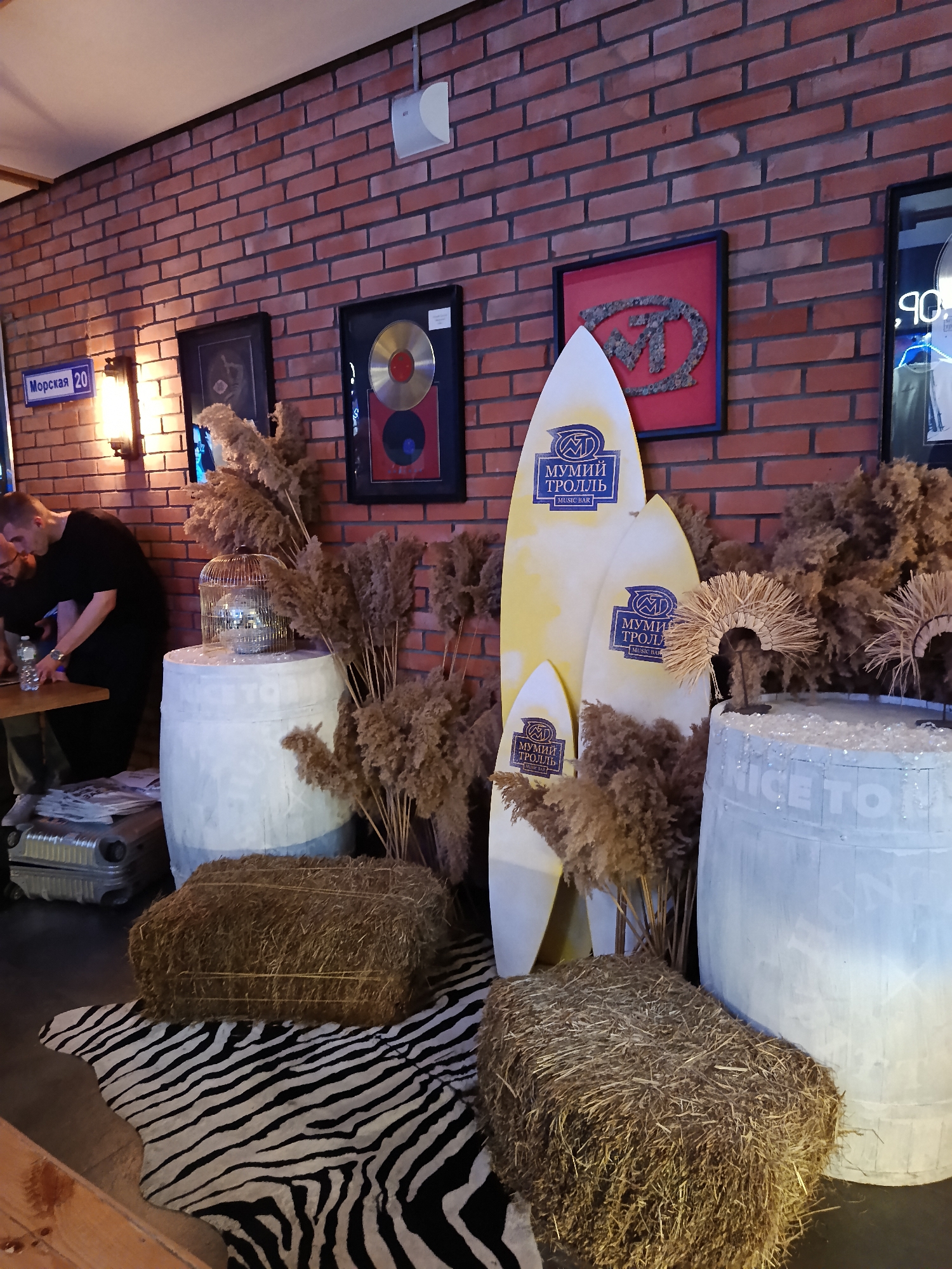 The image shows a decorated interior, likely a themed bar or lounge. The location can be inferred as Moscow, Russia, based on the street sign "Морская 20" (Morskaya 20) visible on the wall.
A red brick wall serves as the backdrop, adorned with framed artwork, including a gold record. Several surfboards, branded with the logo "МУМИЙ ТРОЛЛЬ" (Mumiy Troll) and the text "MUSIC BAR," are propped up. The floor is covered with a zebra-patterned rug, upon which hay bales are placed. Two white-painted barrels serve as stands for decorative elements: one with a silver birdcage, the other with straw headwear. Behind a table, two people, partially obscured, are visible. Luggage is also present, indicating the venue may be near a travel hub.