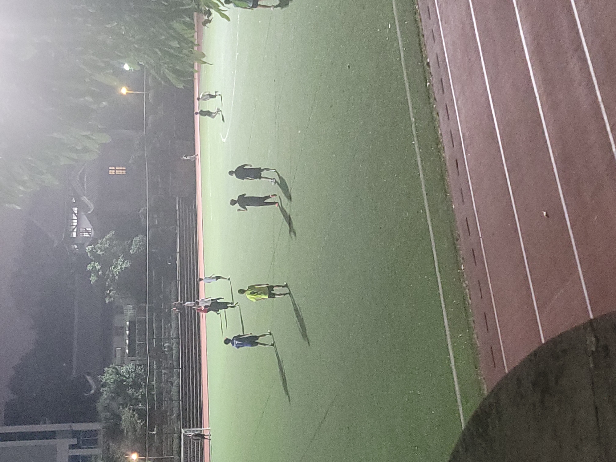 The image shows an outdoor field in Shapingba District, China at night. The field appears to be an artificial turf surface, with a running track adjacent to it. There are approximately 10-12 people on the field, seemingly engaged in a sports activity. Their clothing varies, including light and dark colored athletic wear. The lighting is dim, with artificial light sources casting long shadows. Buildings and trees are visible in the background, partially obscured by the darkness.