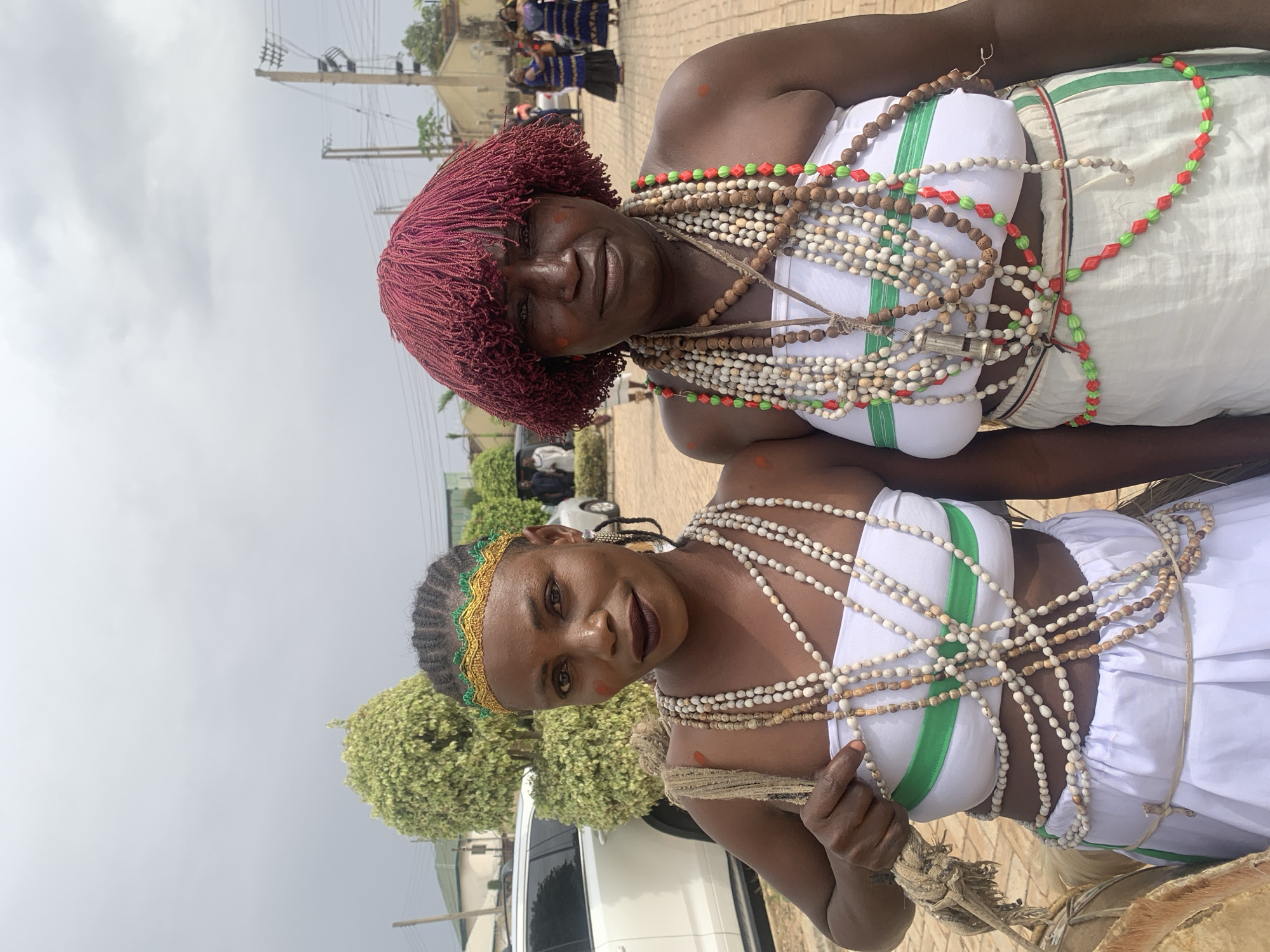Two women are standing closely together outdoors. The woman in the foreground has black braided hair with a green and gold headband. She wears a white garment with a green stripe and many strands of white and brown beads around her neck and torso. The woman behind her wears a similar white garment with a red stripe, strands of red and green beads, and a short red wig. The background includes more people, trees, buildings, power lines, and vehicles. The ground appears to be paved. The location is Jos, Nigeria.