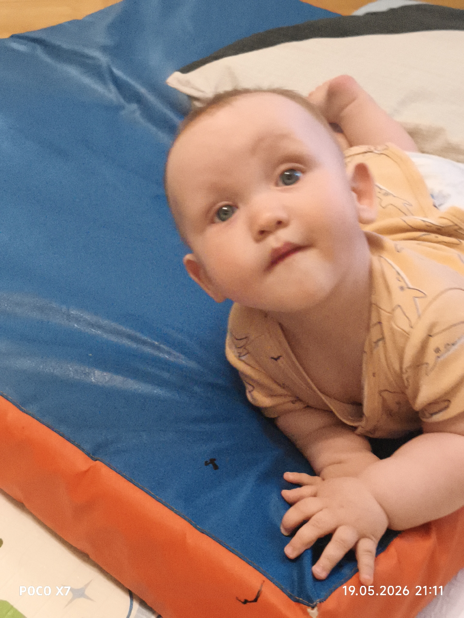 A baby is lying on a blue and orange mat. The baby has fair skin, blue eyes, and is wearing a yellow shirt. The baby is looking up at the camera. There is also a pillow visible behind the baby's head. The location is Unknown, Unknown.