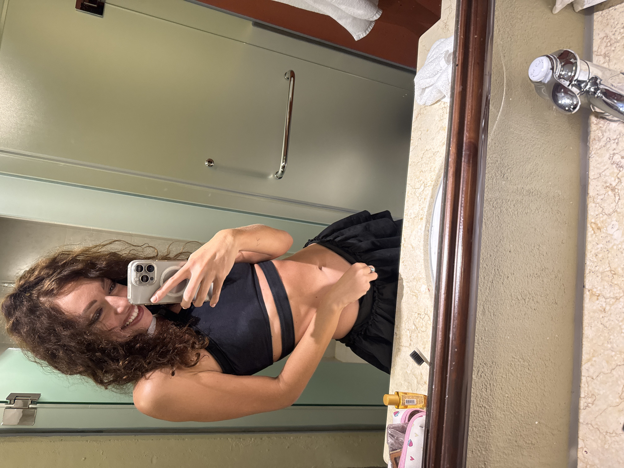 A woman with long, curly brown hair and a bright smile is captured in a mirror selfie within what appears to be a hotel bathroom. She is wearing a black two-piece outfit, comprising a cut-out crop top and a ruffled skirt or shorts. She holds a silver-colored smartphone with multiple cameras, likely an iPhone, in her right hand to take the picture. A ring is visible on her left hand, and a small mark is on her left forearm.
The bathroom features a light-colored marble or stone vanity with a chrome faucet. The faucet handle clearly shows the text "Hot Cold." A wooden trim runs along the mirror or vanity. To the woman's left (in the reflection), a glass shower enclosure with a chrome handle is visible. On the vanity, several items are present, including a yellow bottle (possibly lotion or oil), a toothbrush, and a pink and white patterned item, possibly a cosmetic bag or slippers, along with small white towels. The background behind the shower enclosure appears to be a reddish-brown wall.
The woman's wide smile conveys a happy or content emotion. The image itself is rotated 90 degrees clockwise. The setting suggests an indoor environment, typical of a hotel. Based on the provided context, the location is in Đà Nẵng, Vietnam.