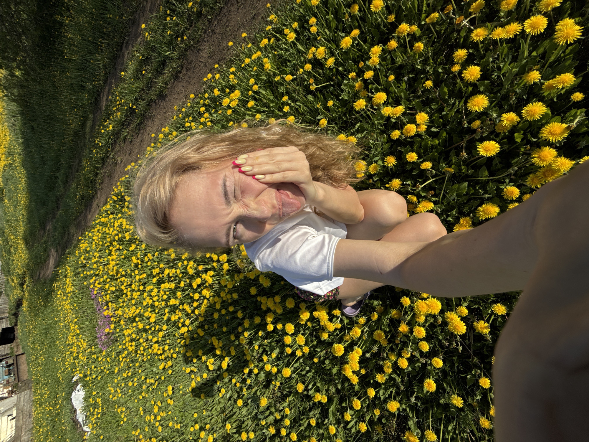 A woman is crouching in a field of yellow dandelions, appearing to take a selfie. She is wearing a white t-shirt and a floral patterned skirt, with what appears to be a purple shoe visible. Her hair is blonde and wavy, and she is sticking her tongue out while holding her hand near her face. The location includes a field with a building on the left side of the image, which is consistent with a rural area like Nizhnyaya Chermoda, Russia. The ground is covered in dandelions, and there is some green grass and foliage visible along the edges of the field. The image is taken at an angle, looking down at the woman and the field.