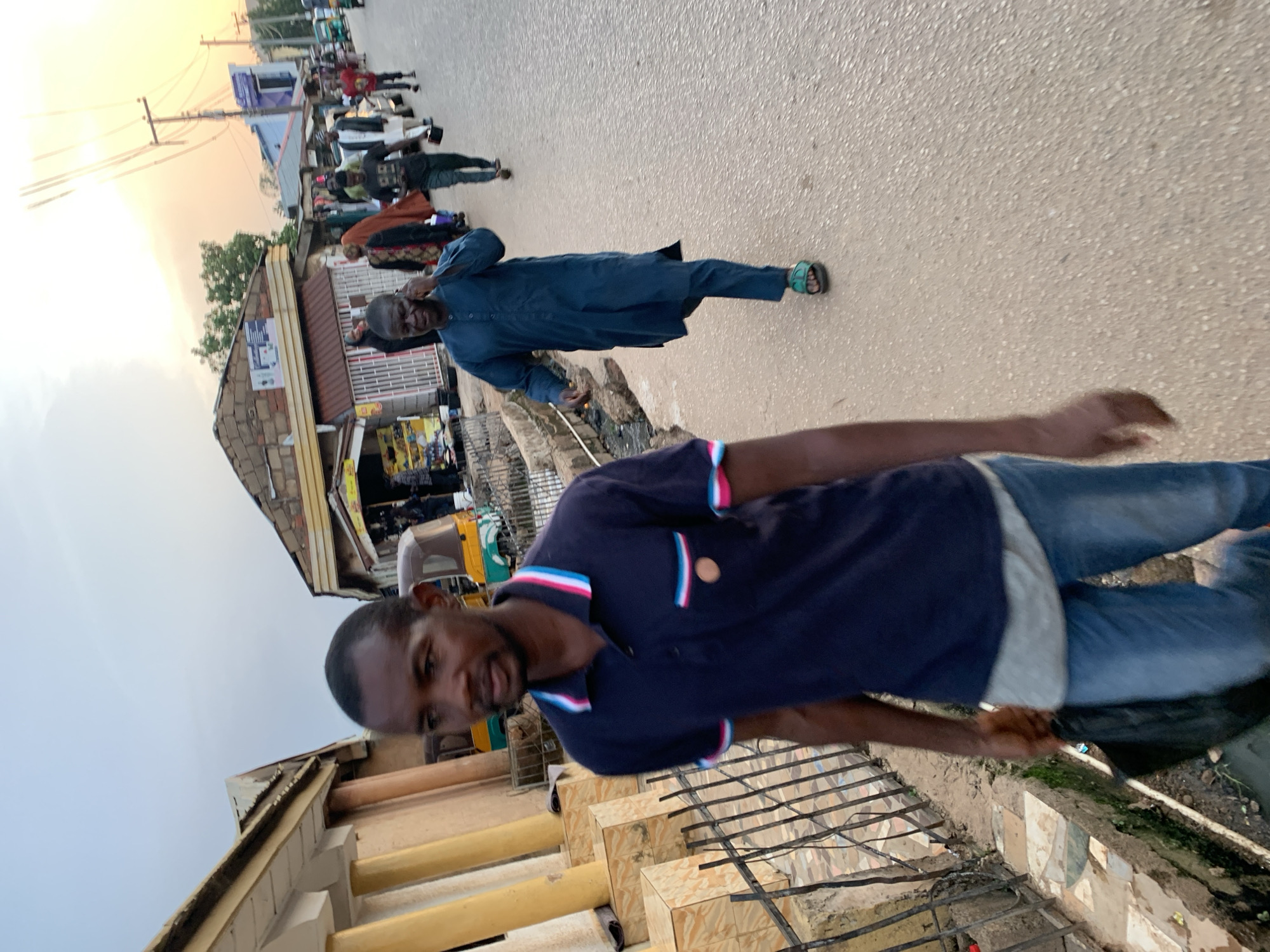 In Gura Top, Nigeria, a street scene unfolds under a pale, hazy sky at dusk. The foreground is dominated by a man walking towards the camera, his dark blue polo shirt featuring striped trim on the collar and pocket, and he is wearing blue jeans and carrying a black plastic bag. Behind him, other individuals traverse the dusty, paved road. To the right, a man in a flowing teal garment, speaking on his mobile phone, walks away from the viewer. Further down the street, a procession of people, some in traditional attire, move amidst scattered shops and vehicles, including a yellow and green auto-rickshaw. The architecture is a mix of simple, weathered buildings, some adorned with awnings and signage. Power lines crisscross the sky, hinting at the urban environment.